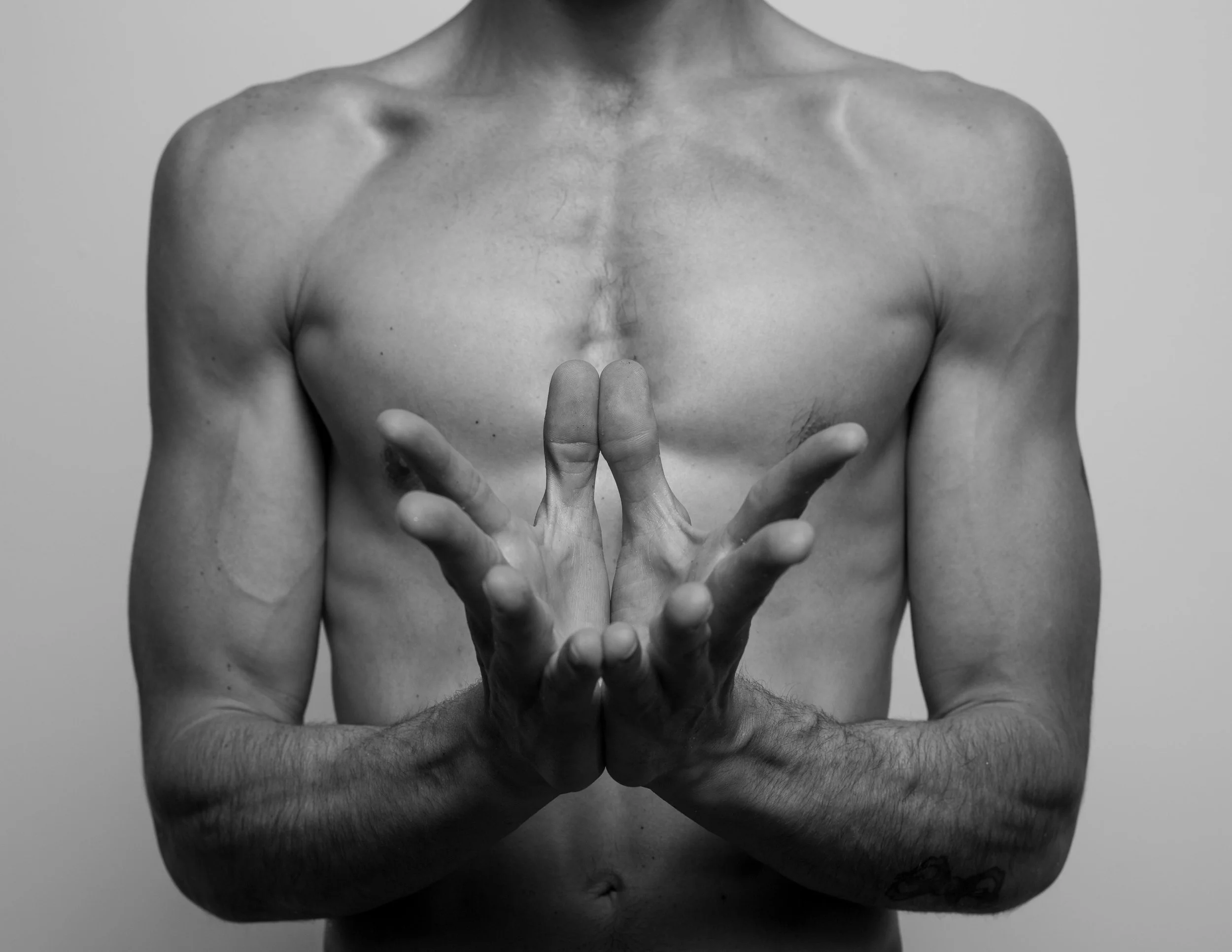 Black and white photo of a topless man with tattooed arms, holding his hands together in a prayer-like gesture.