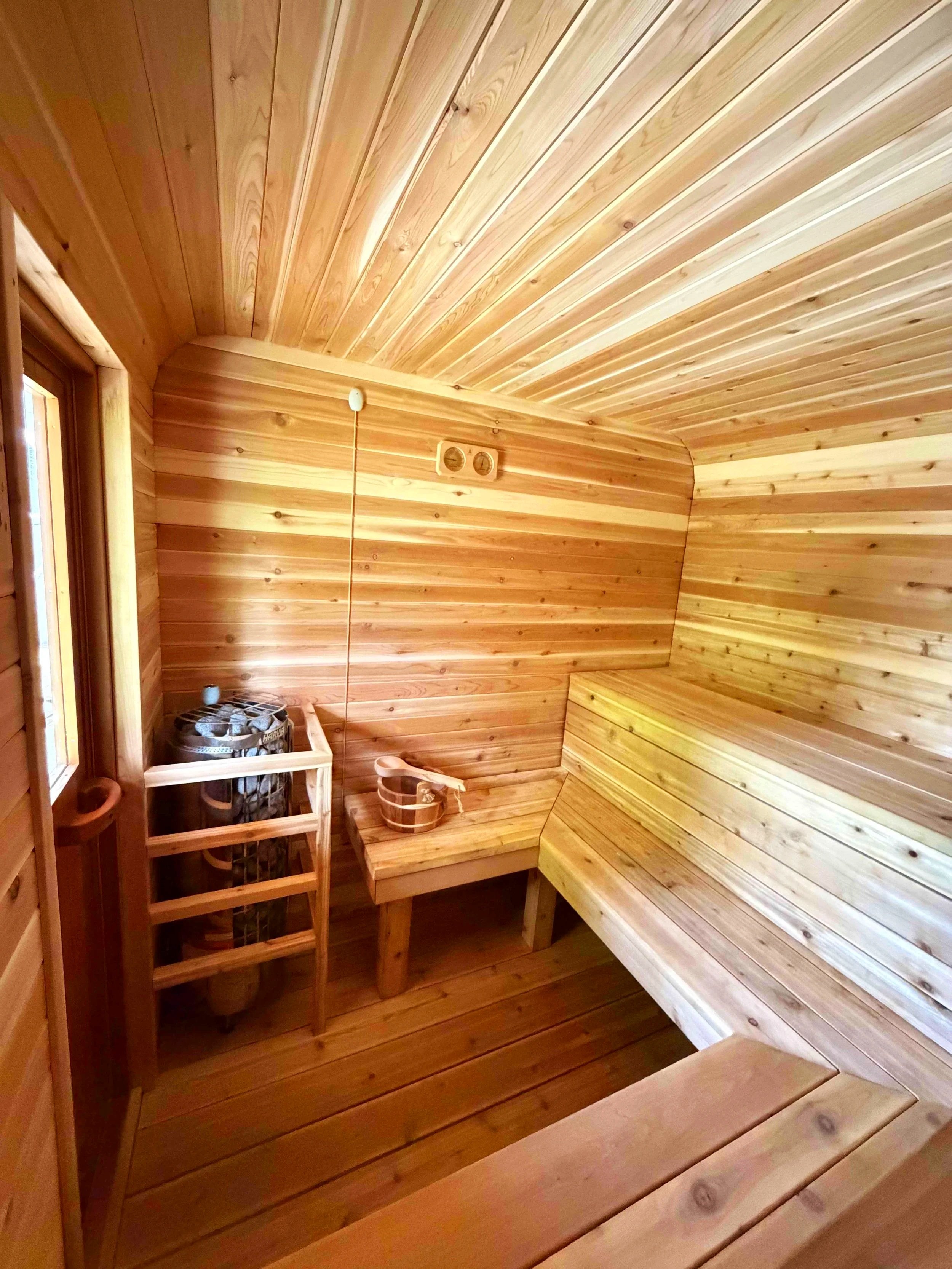 A wooden sauna room with wooden benches, a small heated stove, and a heater with stones, all made of natural wood.