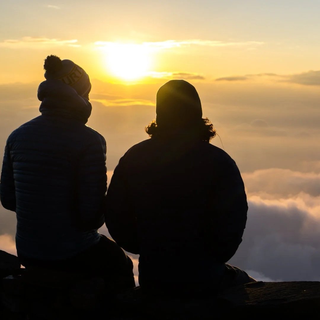 Two people sitting on a rock at sunset, facing the sky with clouds below, enjoying the view.