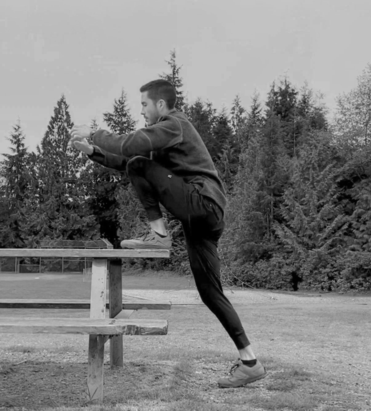 A man doing a stretching exercise outdoors, standing on one leg with the other knee raised and resting on a picnic table, surrounded by trees and greenery.