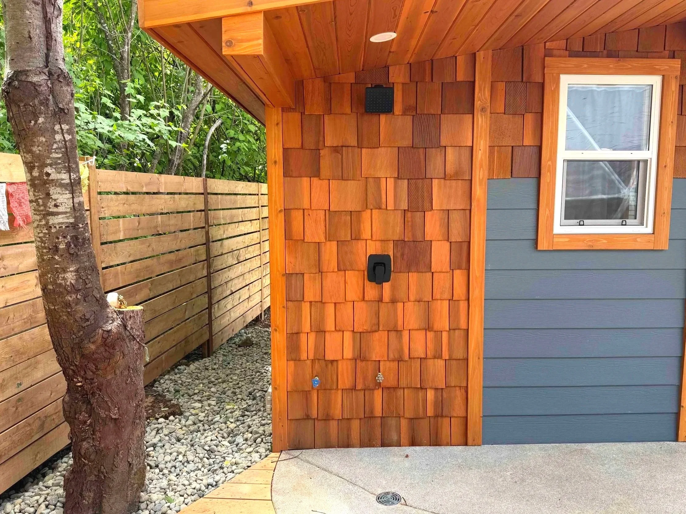 Part of a house exterior with blue siding, a window, and a wooden shingle wall, next to a gravel area and a wooden fence.