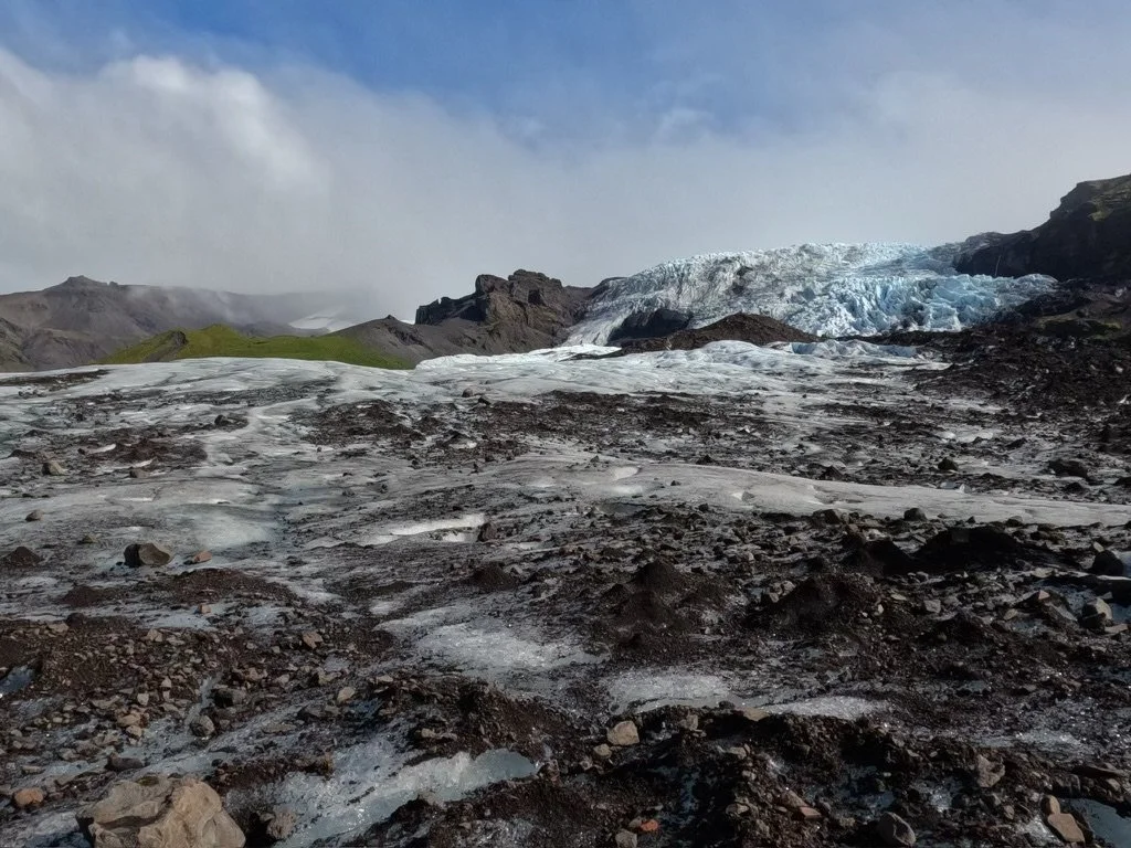 Glacier flowing down a mountain with rocky and moss-covered terrain in the foreground.