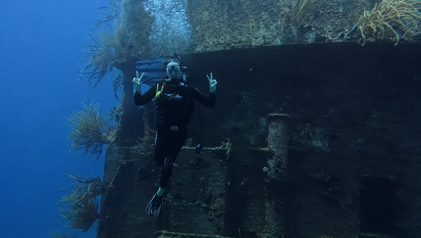 A scuba diver underwater near a sunken shipwreck, making peace signs with both hands.