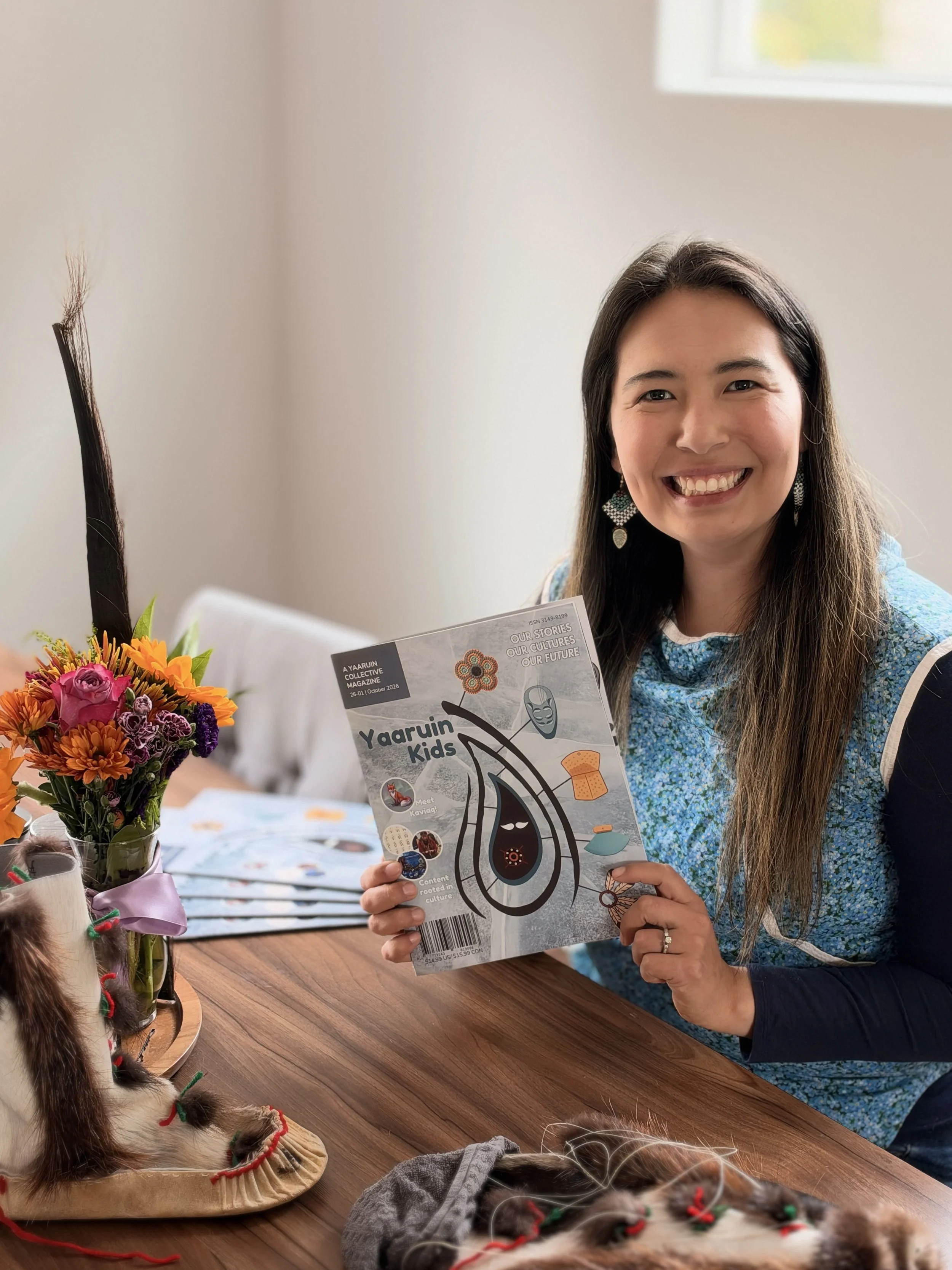 A woman holding the Alaska Native children's magazine Yaaruin Kids while seated at a table.