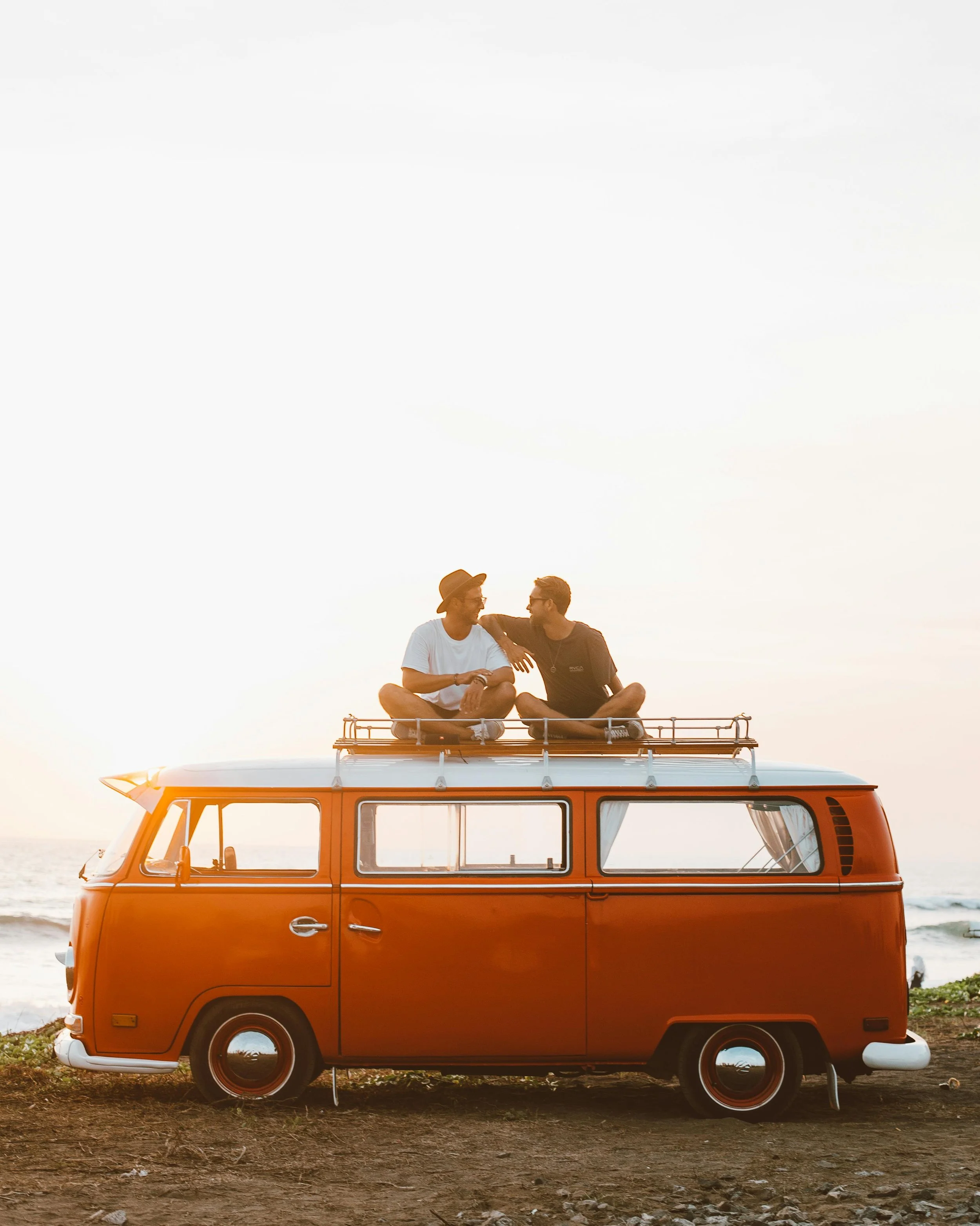 Two men sitting on top of a vintage orange van at the beach during sunset, engaging in conversation.