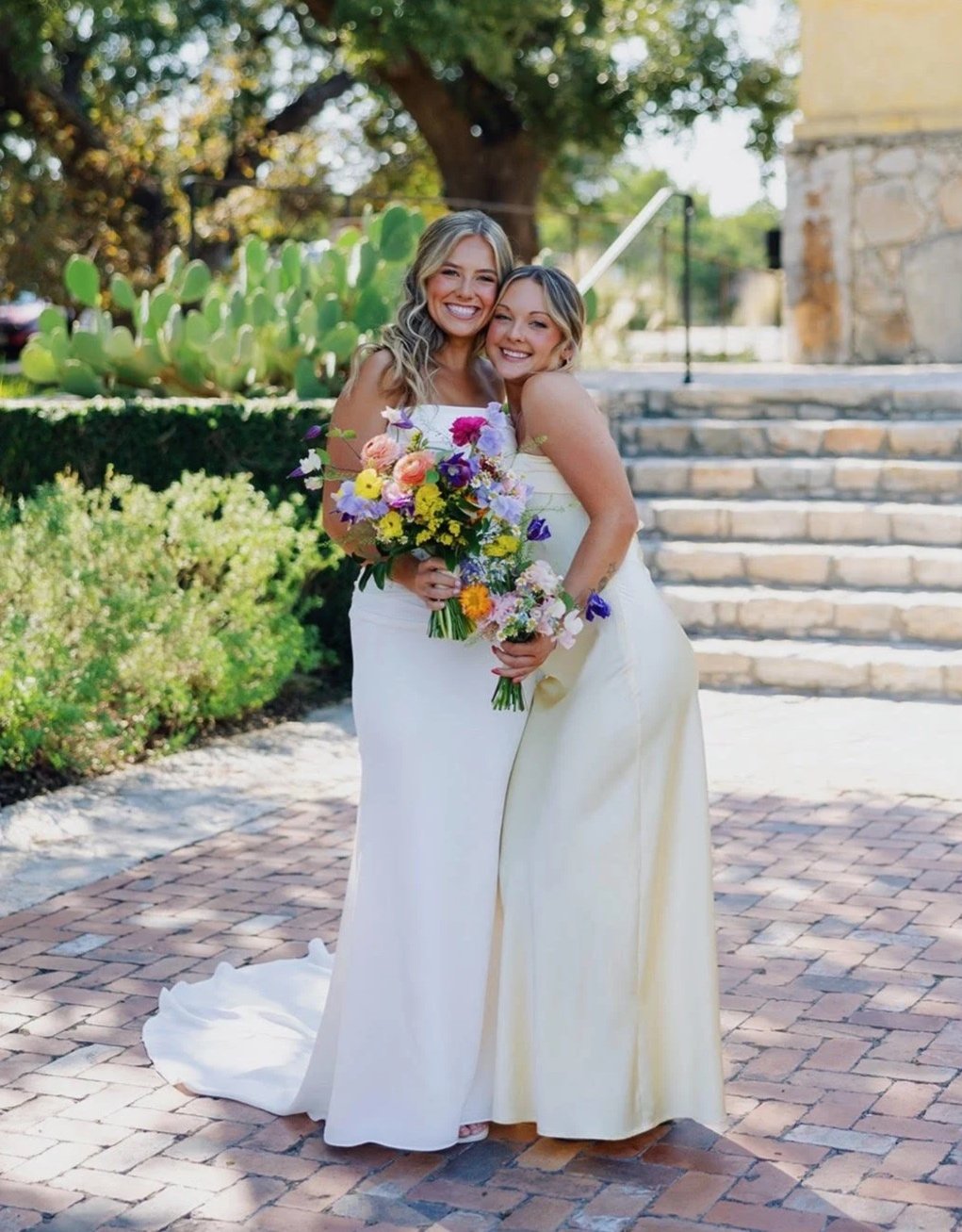 Two women in wedding dresses hugging outdoors on a brick pathway, holding colorful bouquets of flowers. There are green plants and a large cactus in the background.