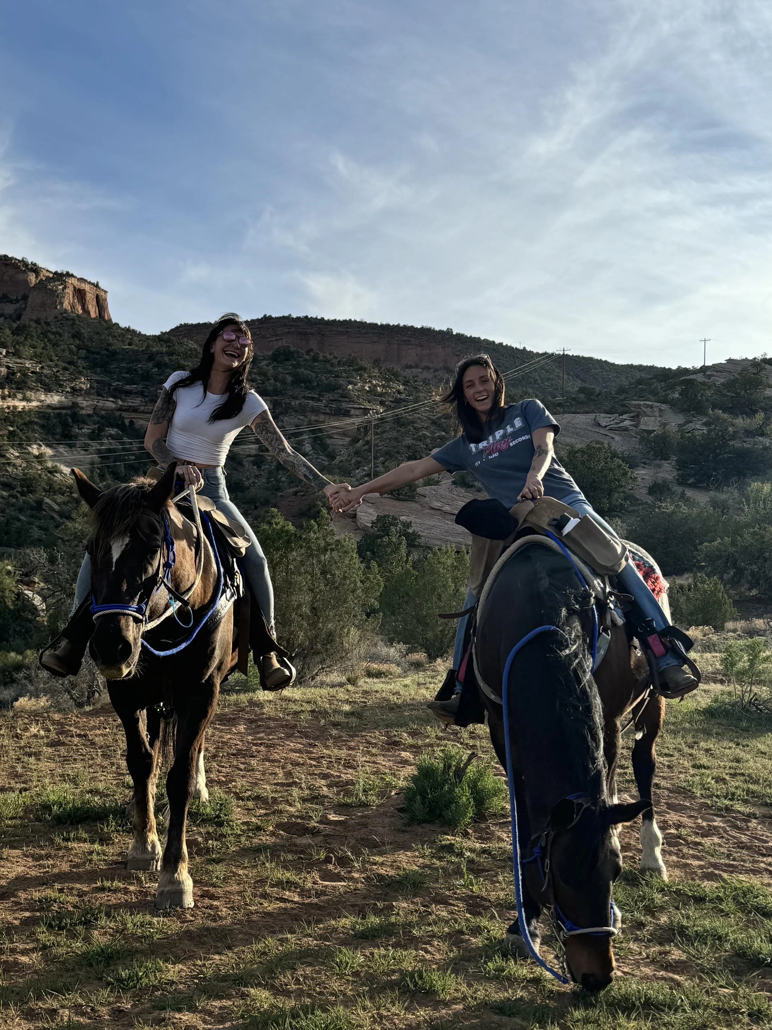 Two women riding horses, holding hands, in a scenic outdoor landscape with hills and trees, smiling and enjoying the moment.