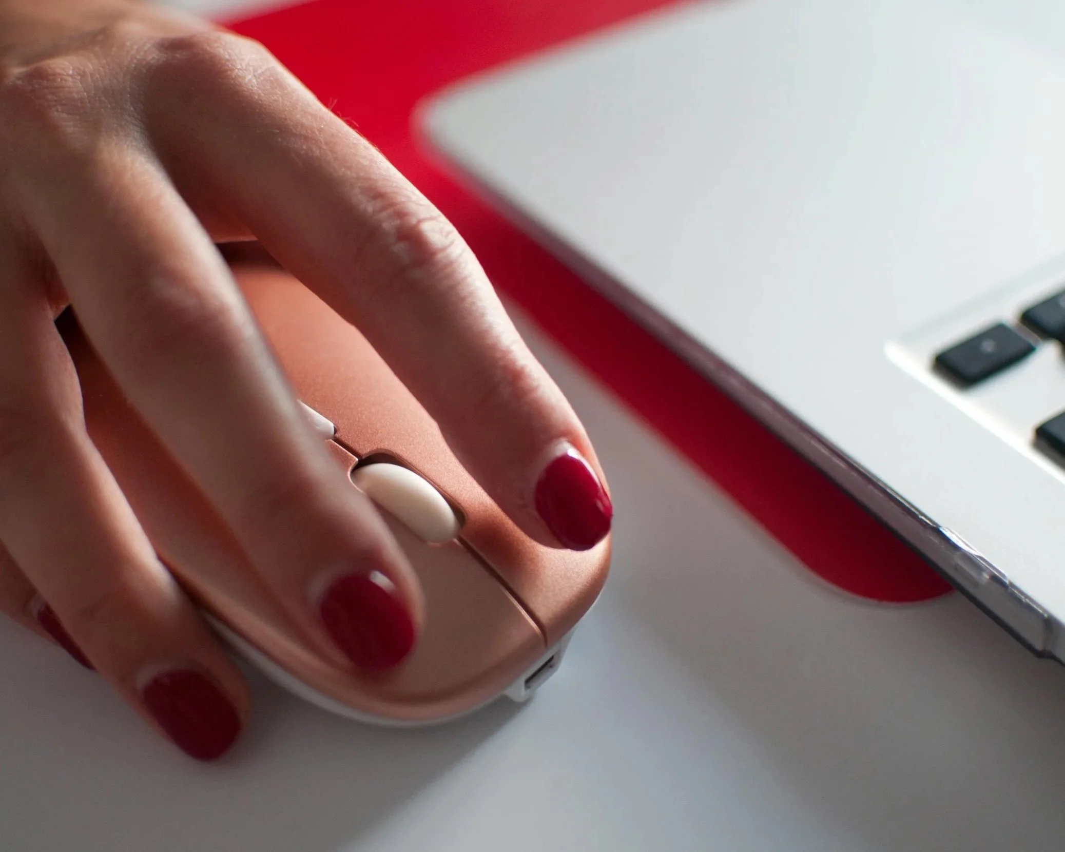 Close-up of a person's hand with red painted nails using a white computer mouse on a desk, with a laptop partially visible.