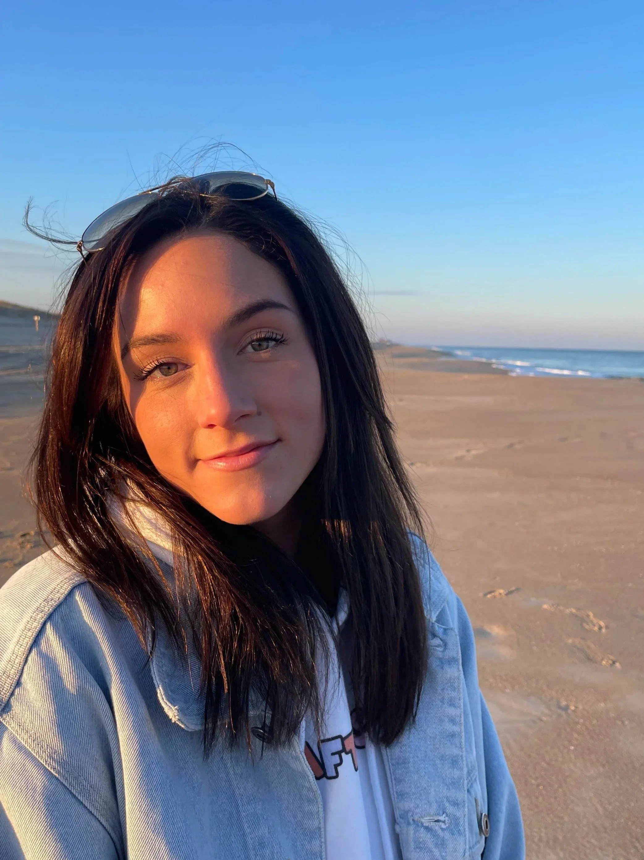 A young woman with dark hair and a denim jacket standing on a sandy beach during sunset.