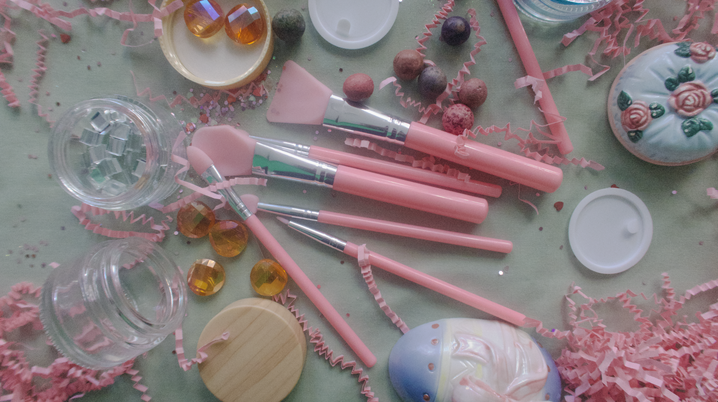 Party decoration with pink makeup brushes, plastic confetti, small jars, colorful beads, a painted ceramic bunny figurine, a cupcake-shaped ornament, and shredded pink paper.