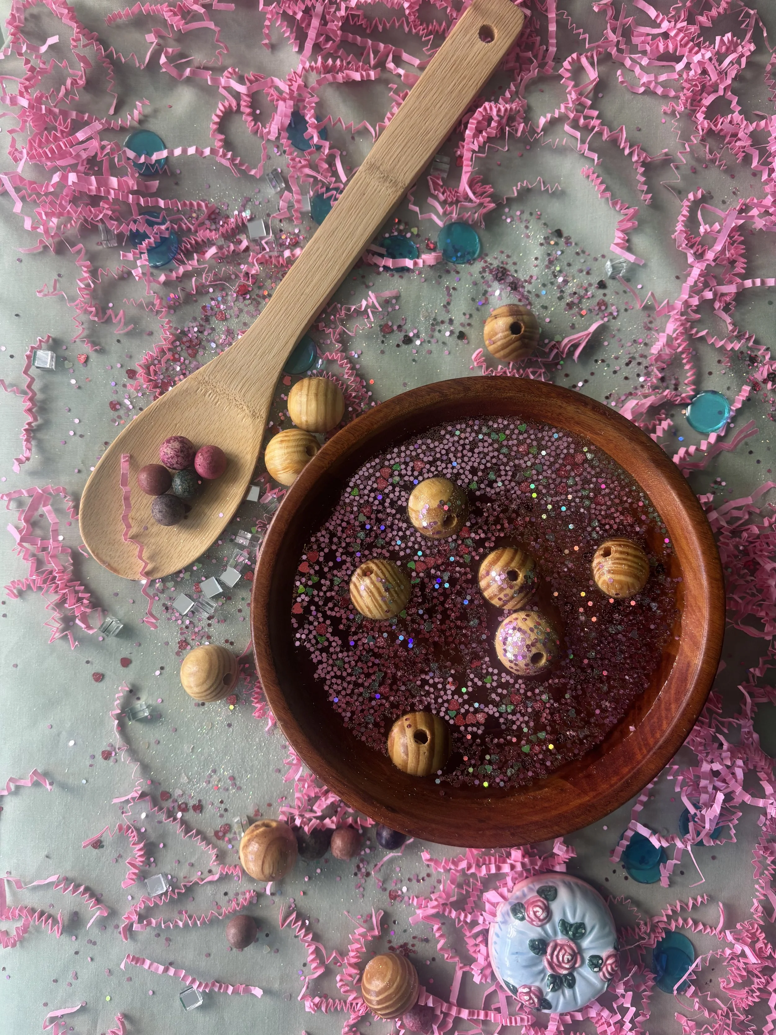 A wooden bowl filled with water and pink glitter, holding seven round wooden beads with striped patterns. Pink streamers and shiny confetti are scattered on a pastel surface, with additional beads, a wooden spoon, and a decorated ceramic ball nearby.