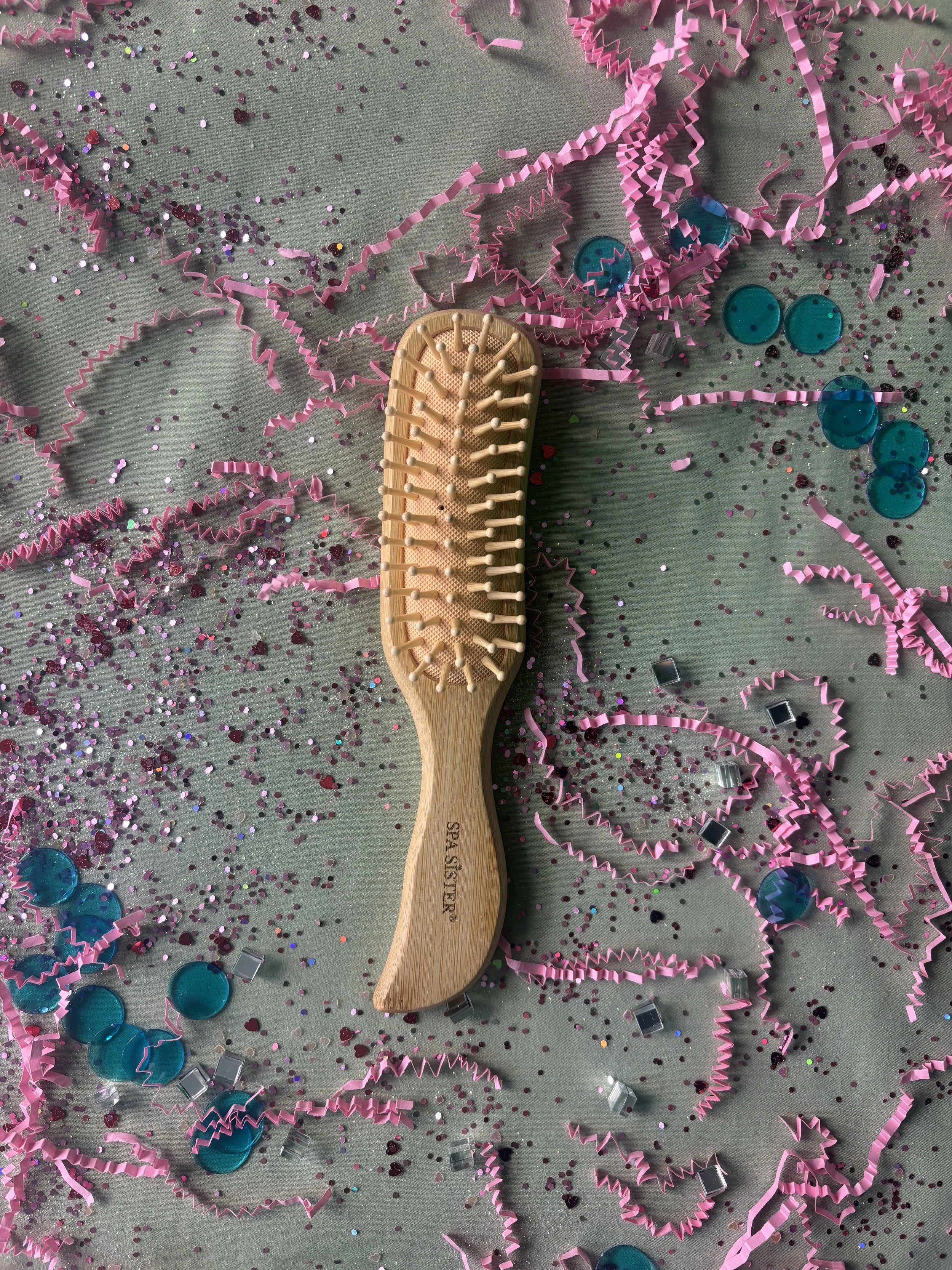 A wooden hairbrush on a surface decorated with pink confetti, pink curly ribbon, and blue bottle caps.