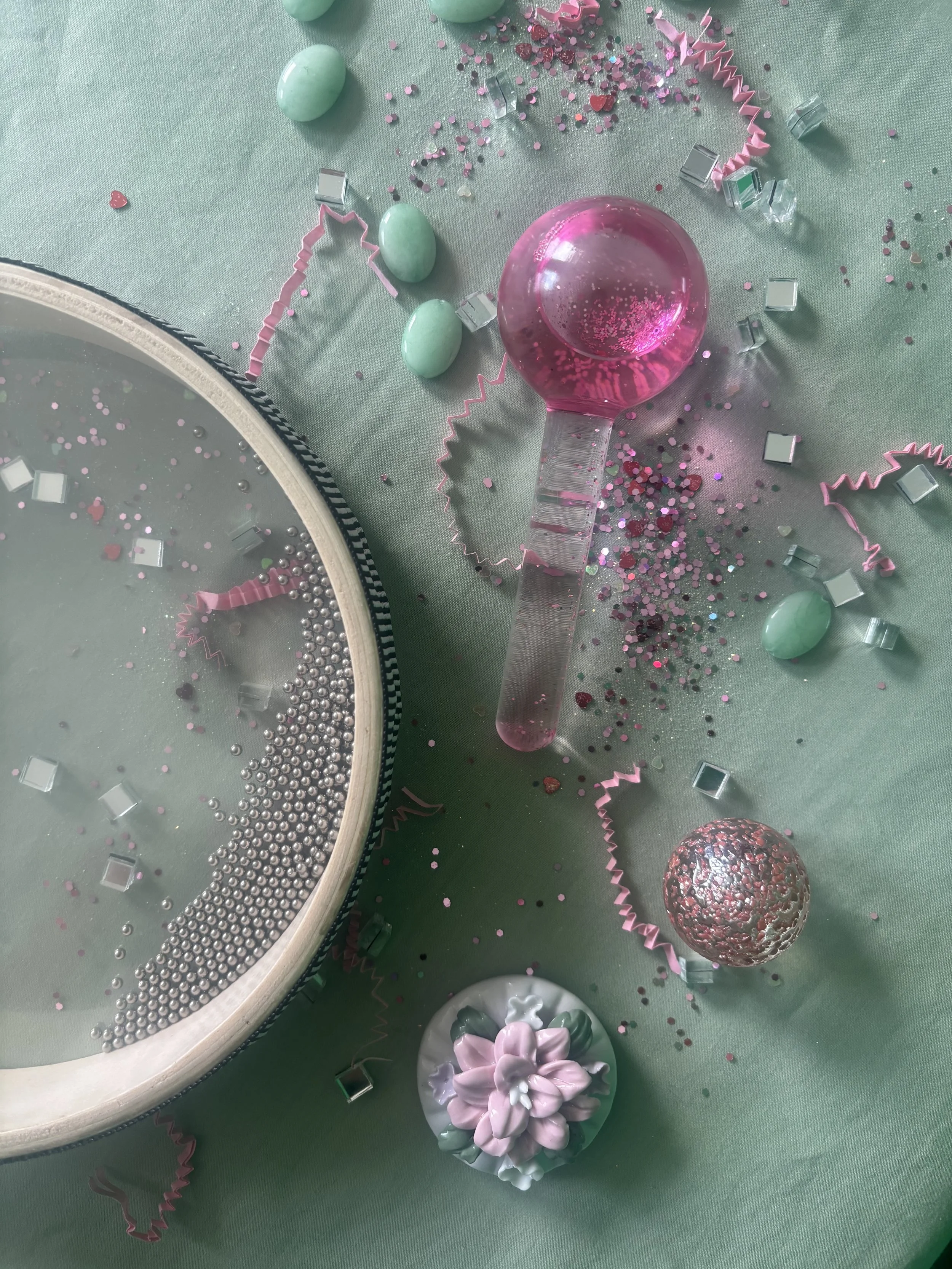 Party decorations on a green tablecloth, including a round mirror, pink and green candies, pink confetti, a pink and clear glass bubble wand, a metallic pink and silver ball, and a floral-shaped soap or ornament.