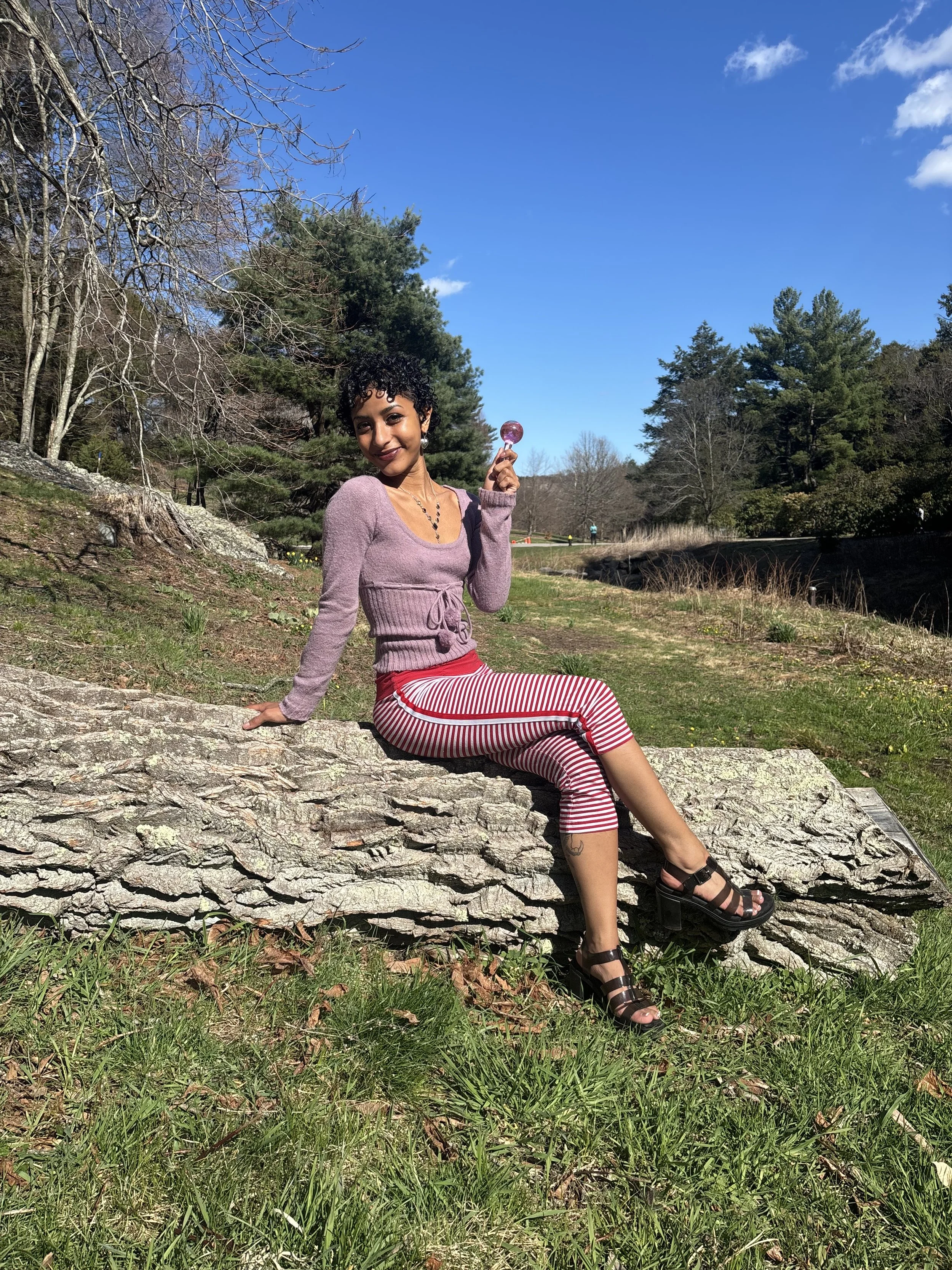 A woman with curly short hair and dark skin sitting on a large fallen tree trunk in a park, holding a purple lollipop, wearing a mauve sweater, red and white striped pants, and black sandals, with a background of trees, grass, and a bright blue sky.