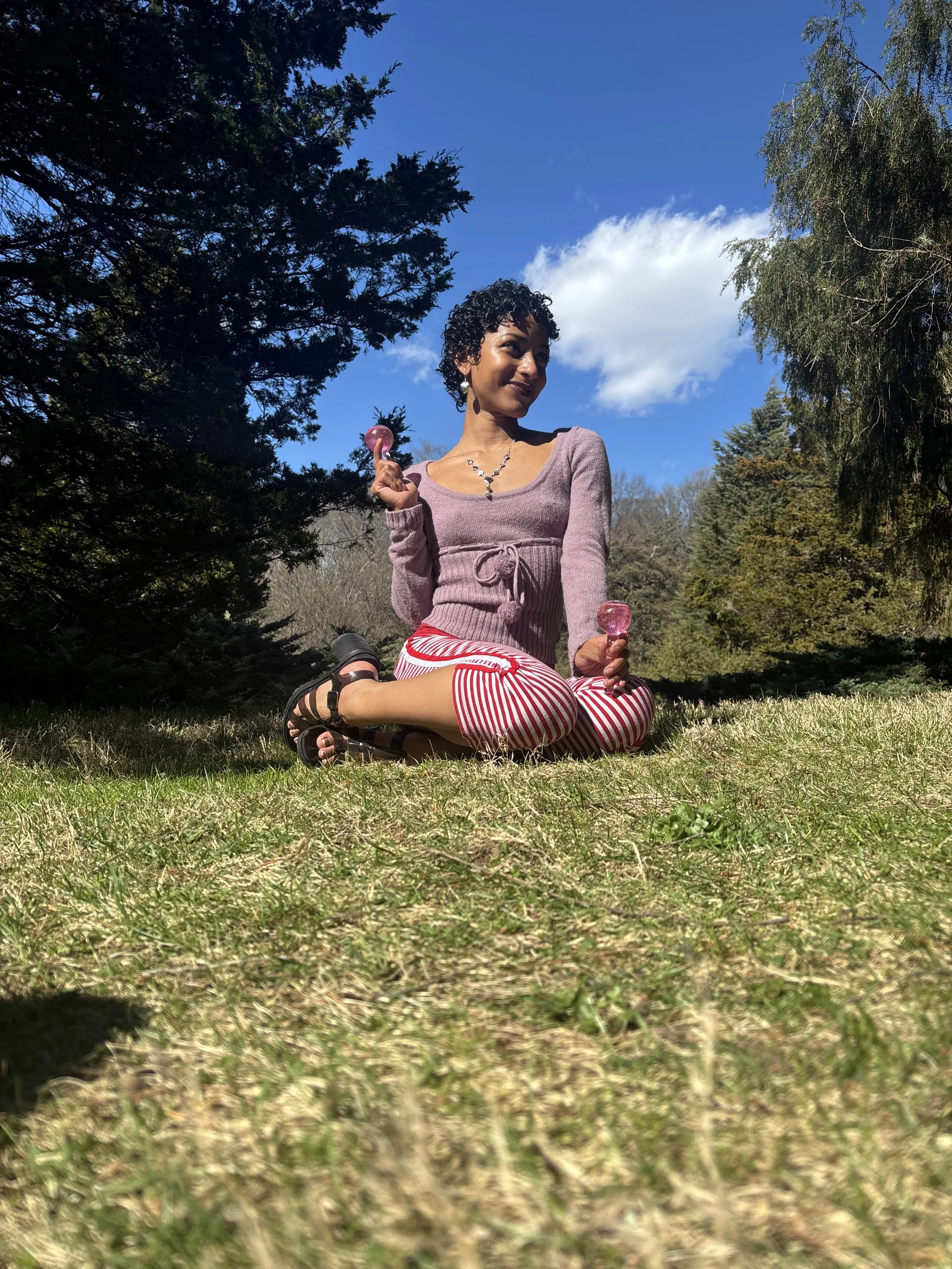 A woman sitting cross-legged on grass in a park, holding pink gumballs, with trees, blue sky, and a few clouds in the background.