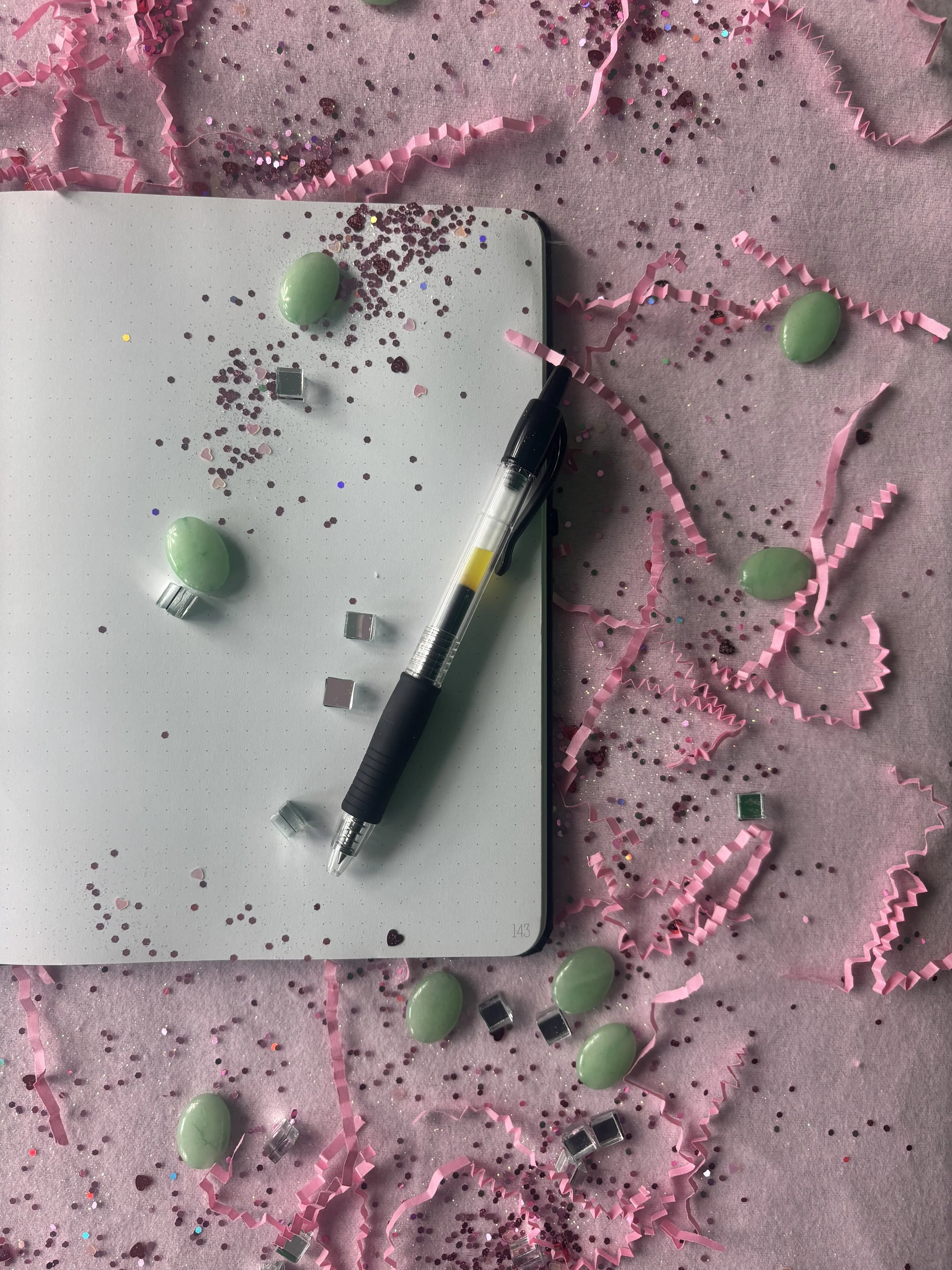 Pink themed party decorations with pink confetti, small square metallic pieces, green oval candies, and a black and clear pen placed on a blank white notepad on pink surface.