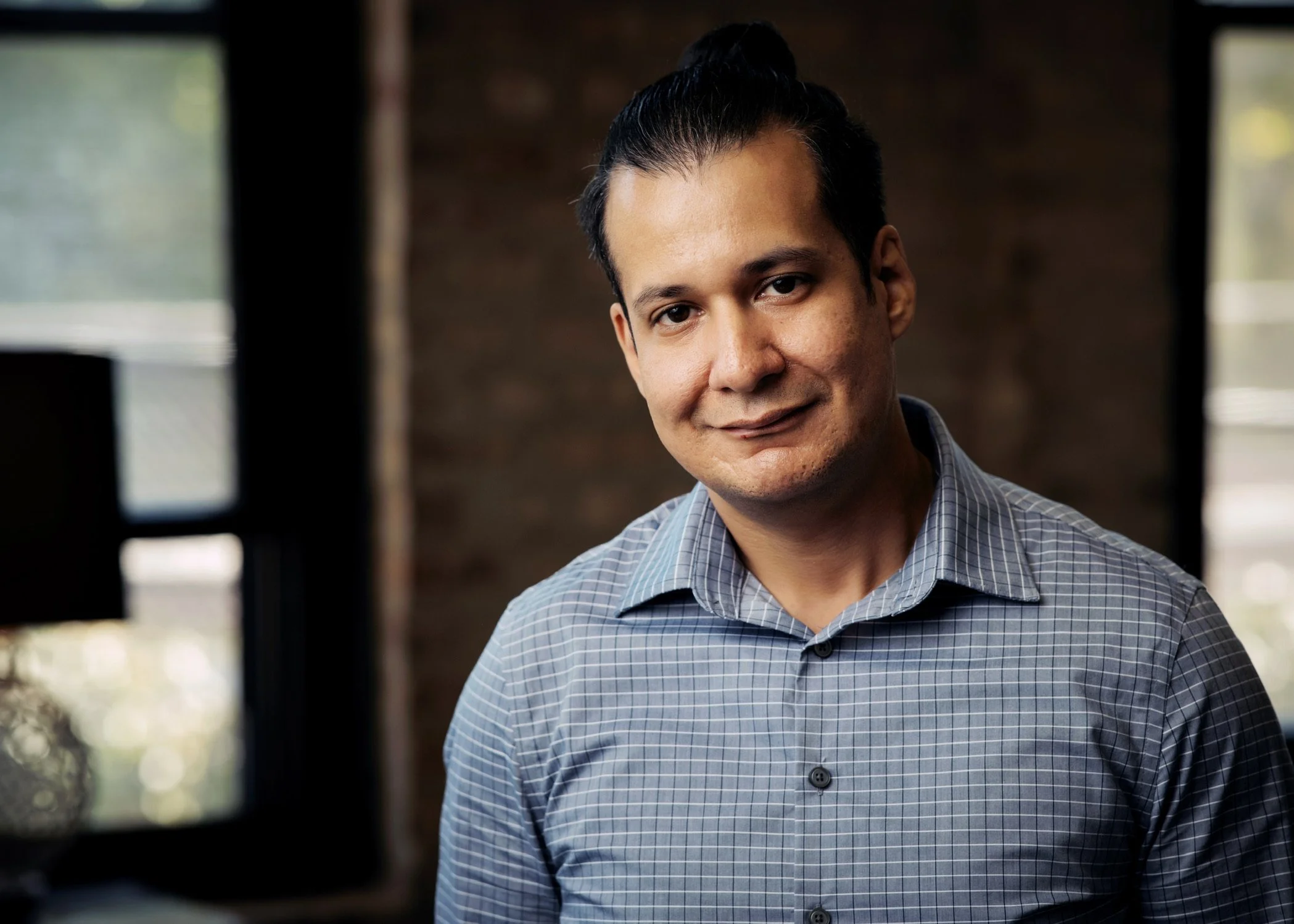 A man with dark hair pulled back, wearing a blue checkered shirt, smiling softly in an indoor setting with natural light coming through windows.