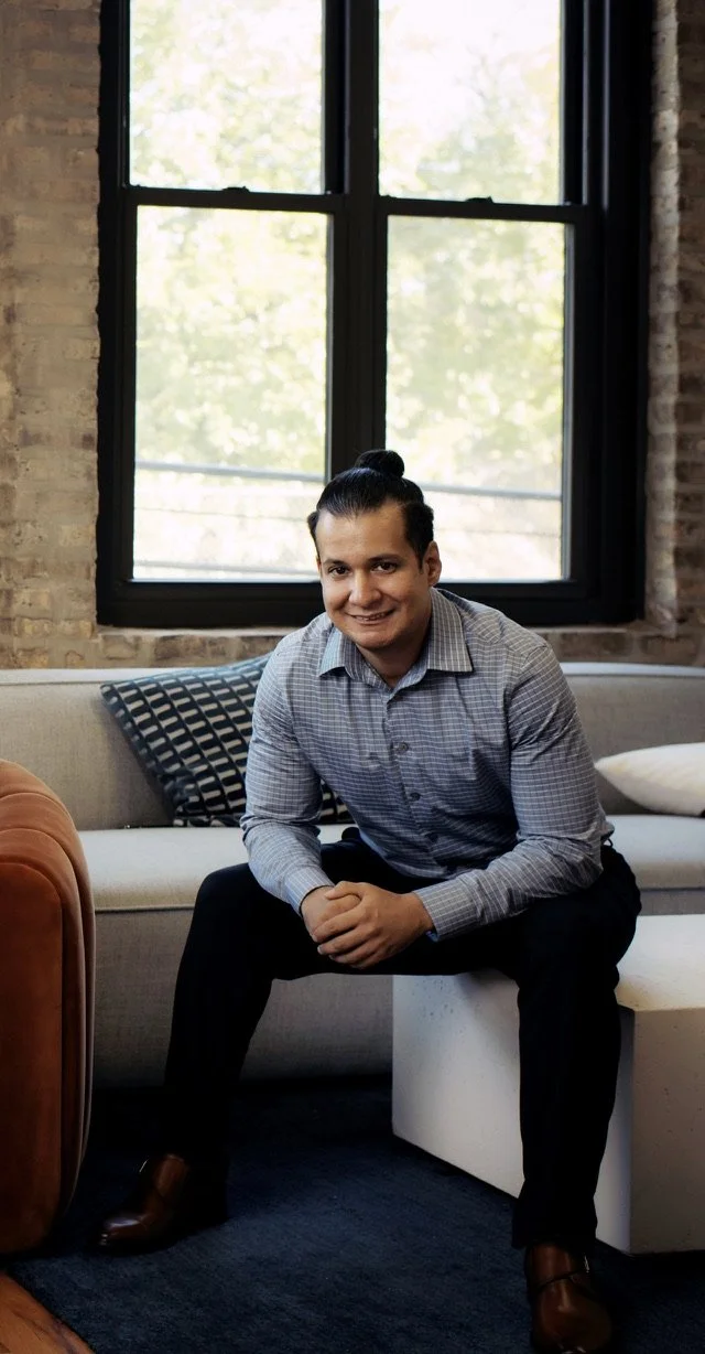 A man sitting on a bench in a room with a large window, brick wall, and patterned pillow behind him, wearing a plaid shirt and dark pants.