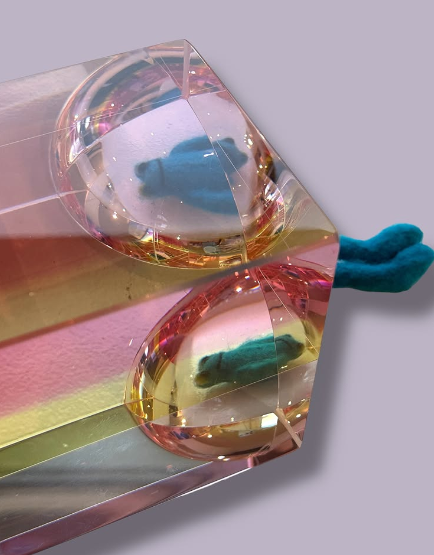 A small plush teal bear inside a clear, faceted gemstone-shaped container, with the bear's reflection visible on the shiny surface.
