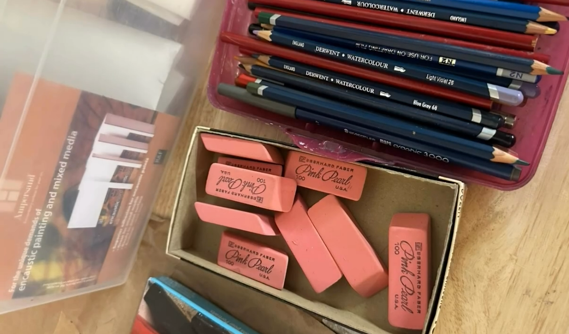 A box of pink erasers next to a container of colored pencils on a wooden surface.
