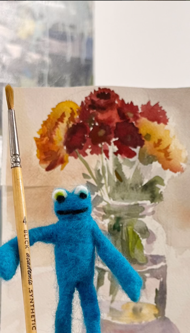 Felt blue puppet with white eyes and black mouth in front of a watercolor painting of a red and yellow flower bouquet in a glass vase, with a paintbrush to the left.