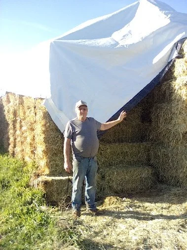 A man standing next to a large hay bale with a tarp covering part of it, outdoors on a farm or rural area.