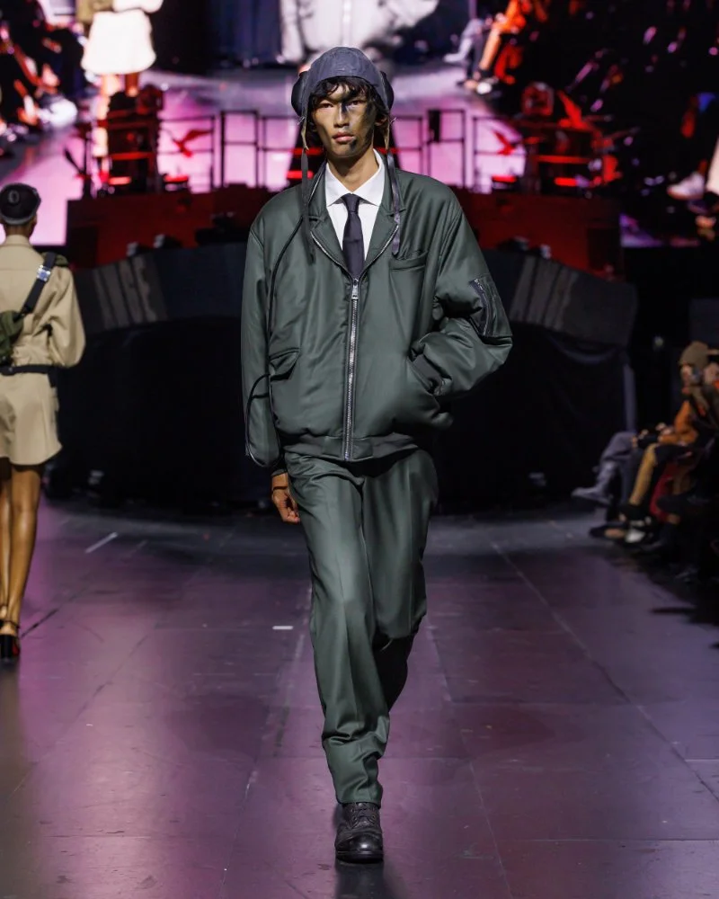 Ensemble vert sombre composé d’une veste zippée et d’un pantalon ample, porté avec chemise, cravate et coiffe ajourée, AWGE SS27.