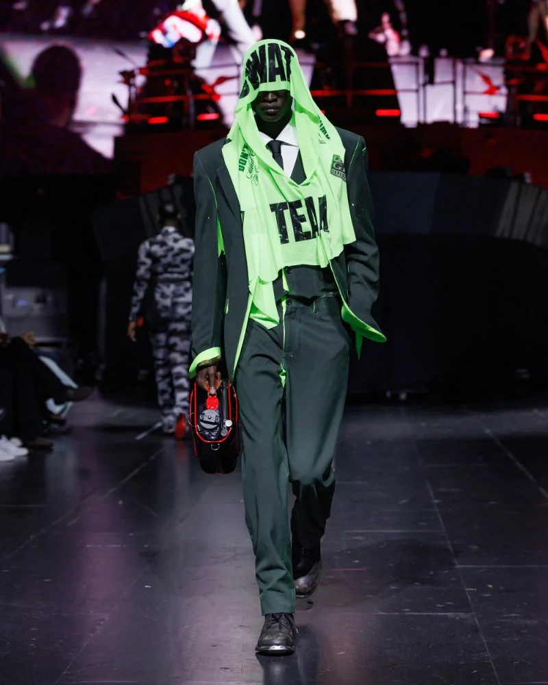 Ensemble de costume vert sombre avec chemise et cravate, sous une étoffe verte fluorescente couvrant la tête et le torse, AWGE SS27.