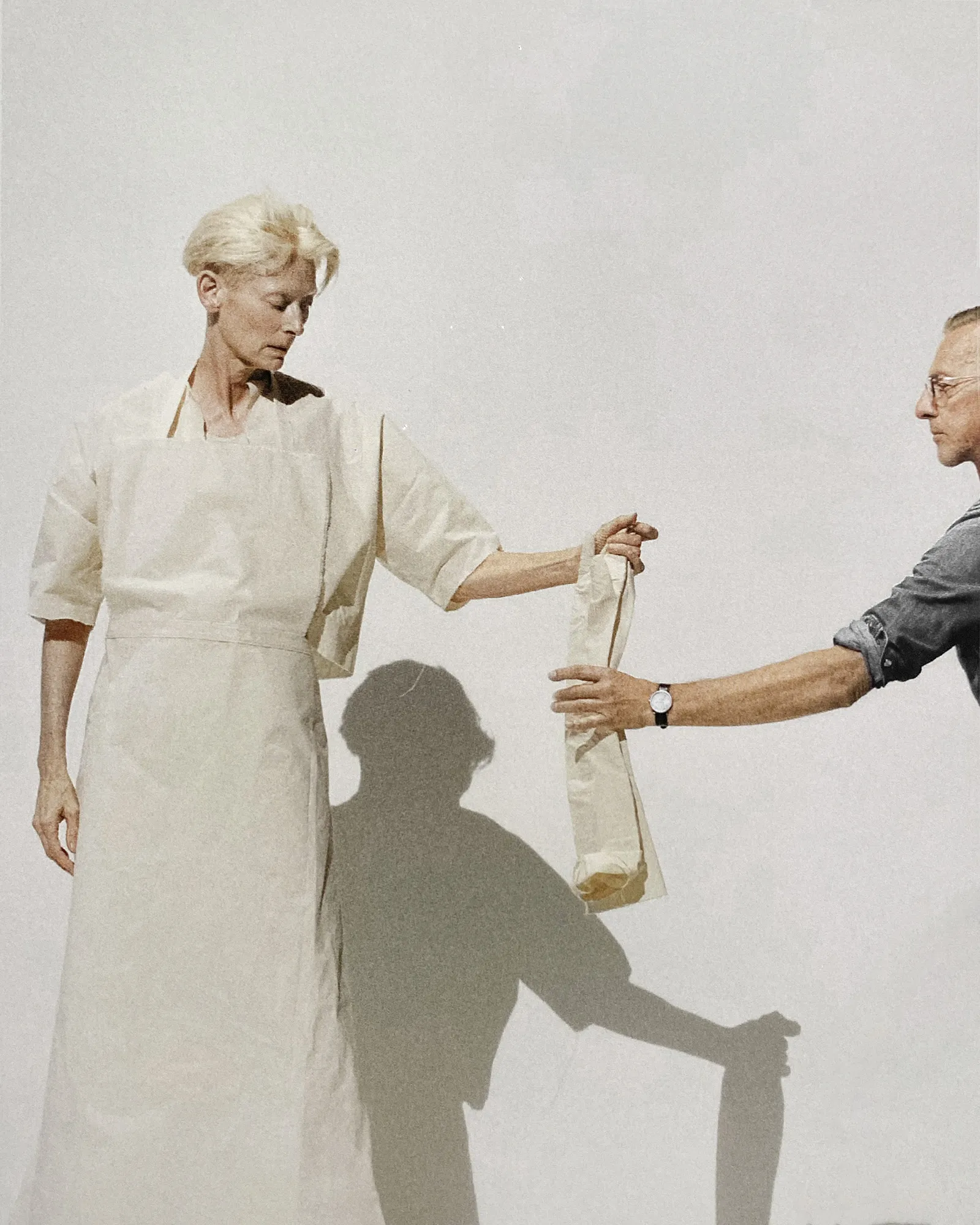 Tilda Swinton en robe blanche tend la main vers Olivier Saillard, leurs ombres projetées sur un fond clair.