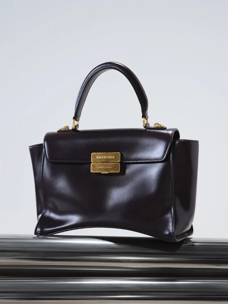 Sac Balenciaga Hourglass Avenue en cuir coloris raisin, posé de trois quarts sur une surface sombre.