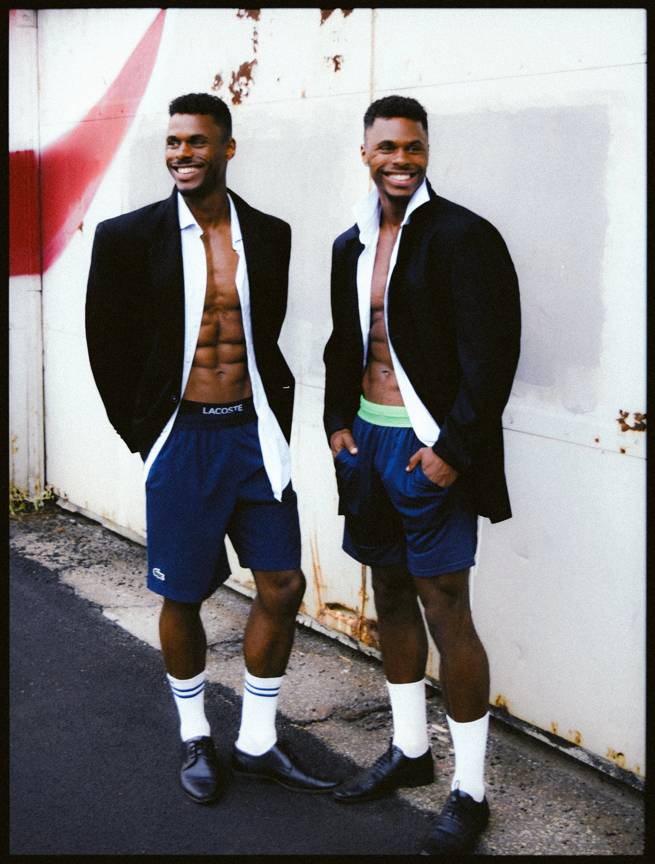 Charles et Maxence Bertimon en vestes sombres et shorts bleus, devant un mur blanc marqué de rouge.