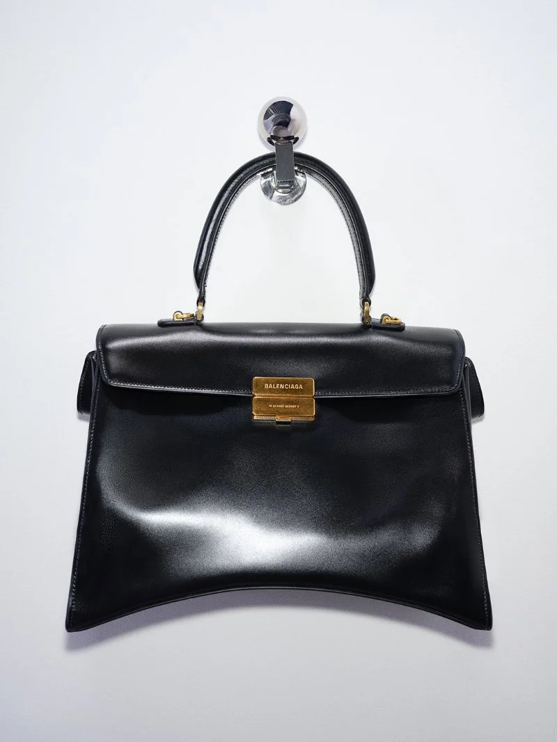 Vue de face du sac Balenciaga Hourglass Avenue noir, avec sa base incurvée et son fermoir doré gravé.