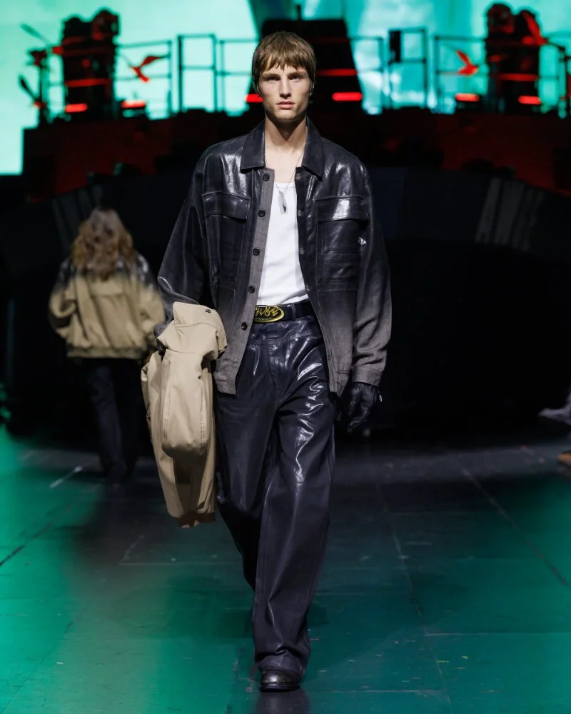 Veste et pantalon noirs brillants avec chemise blanche ouverte, un manteau beige porté au bras, défilé AWGE SS27.
