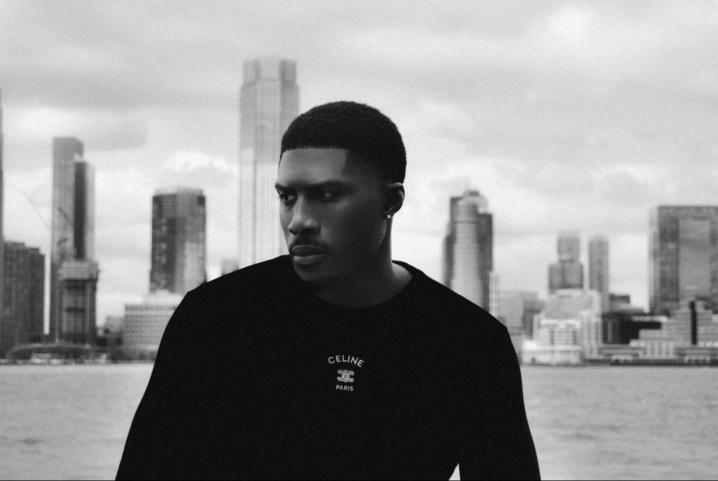 Portrait en noir et blanc d'un homme portant un t-shirt à logo 'CELINE PARIS', avec un panorama urbain de New York en arrière-plan