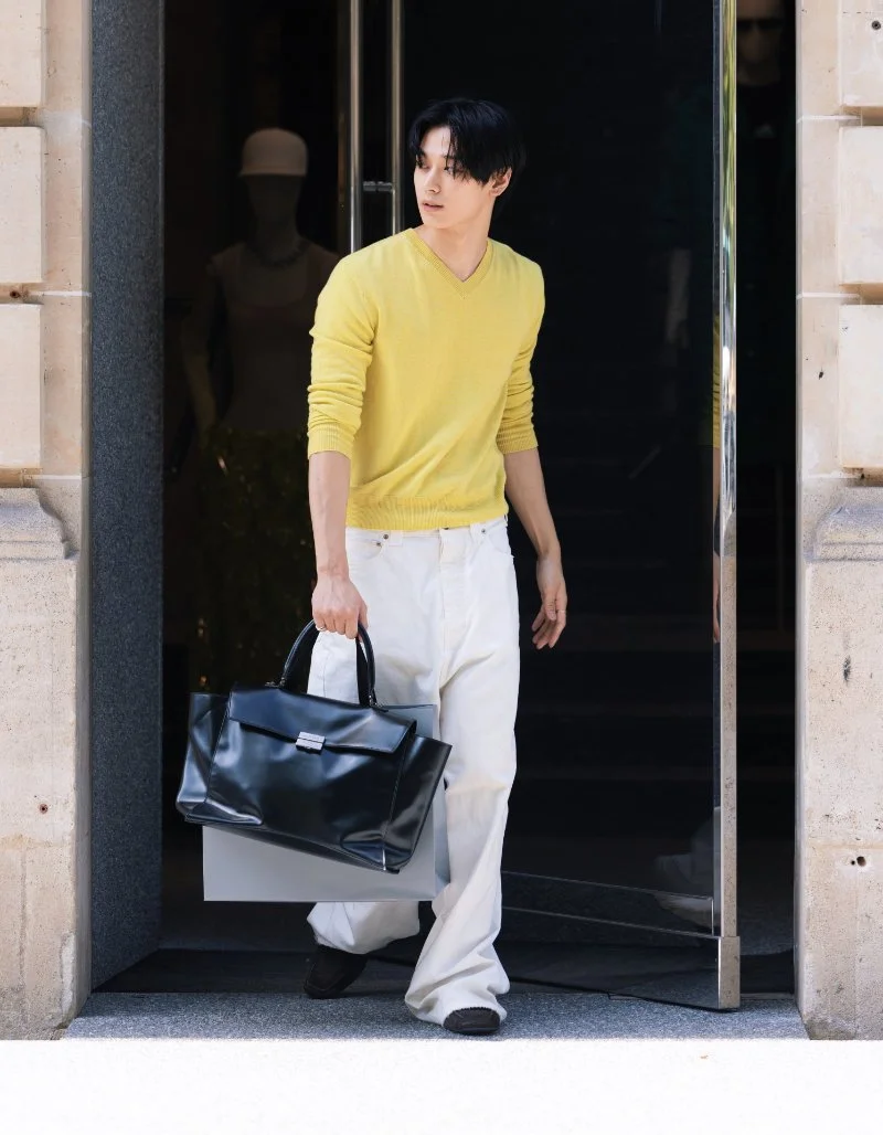 Juyeon sort d’un bâtiment vêtu d’un haut jaune, avec le sac Balenciaga Hourglass Avenue noir à la main.