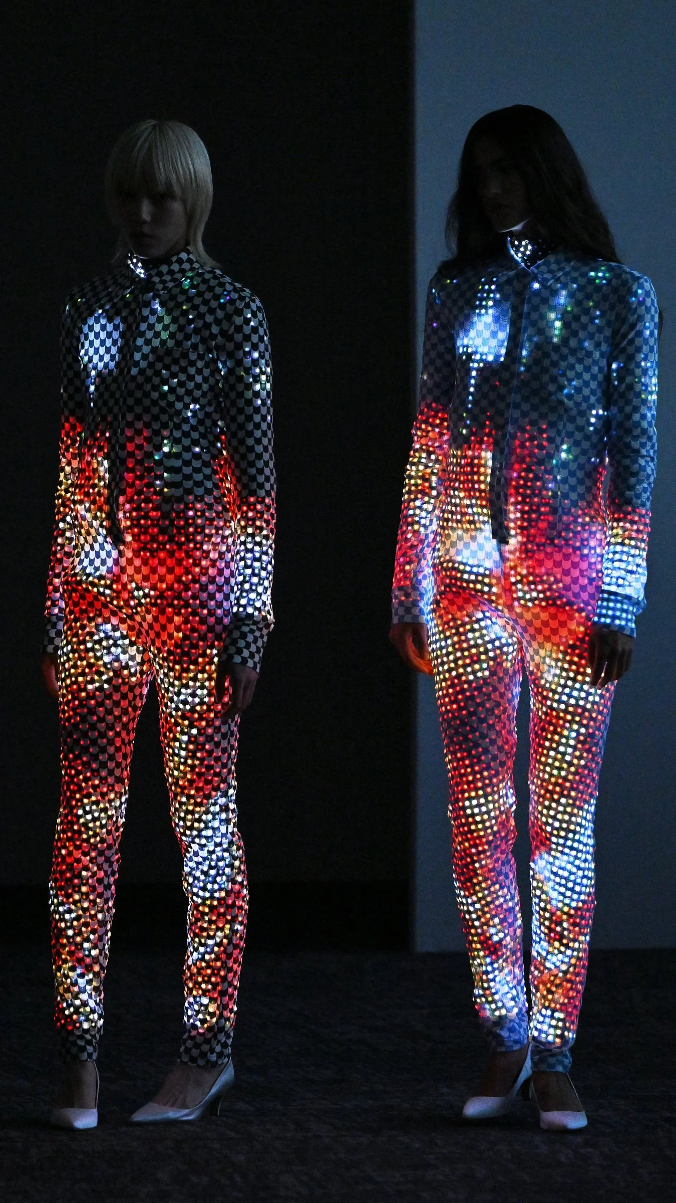 Deux silhouettes aux surfaces lumineuses multicolores se détachent sur un fond sombre lors du final Anrealage printemps-été 2027.