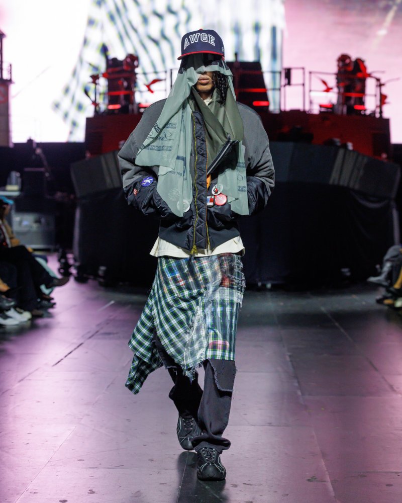 Veste vert grisé, étoffe à carreaux bleus et verts nouée à la taille sur un pantalon sombre, avec casquette et voile, AWGE SS27.