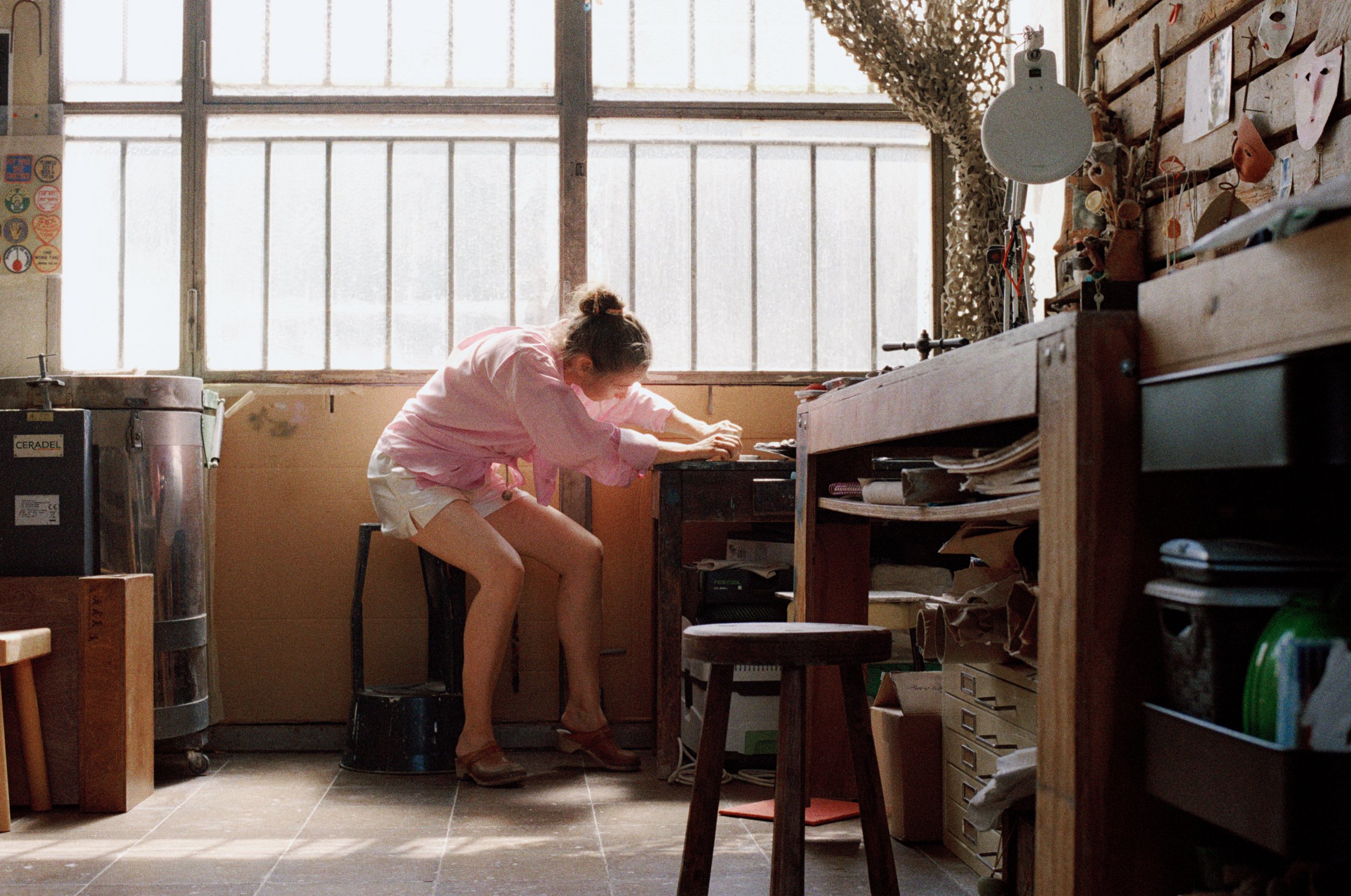 Sylvie Auvray en chemise rose dans son atelier, près d’une table de travail éclairée par de grandes fenêtres.