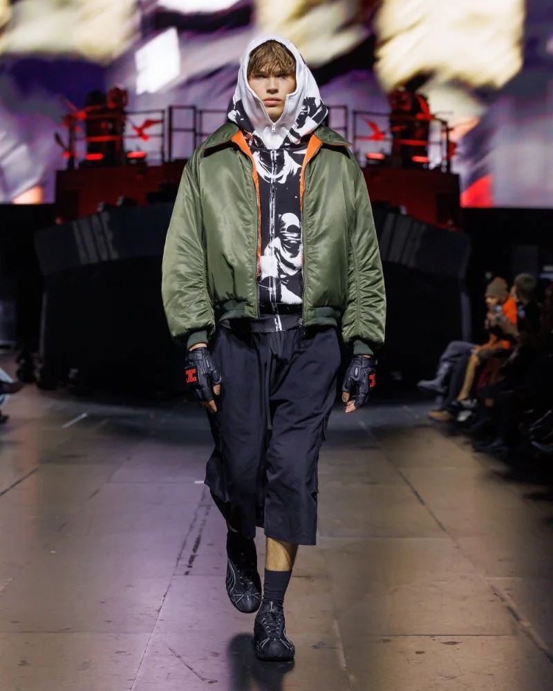 Bomber kaki, haut graphique noir et blanc, pantalon court bleu marine et étoffe imprimée autour de la tête, défilé AWGE SS27.