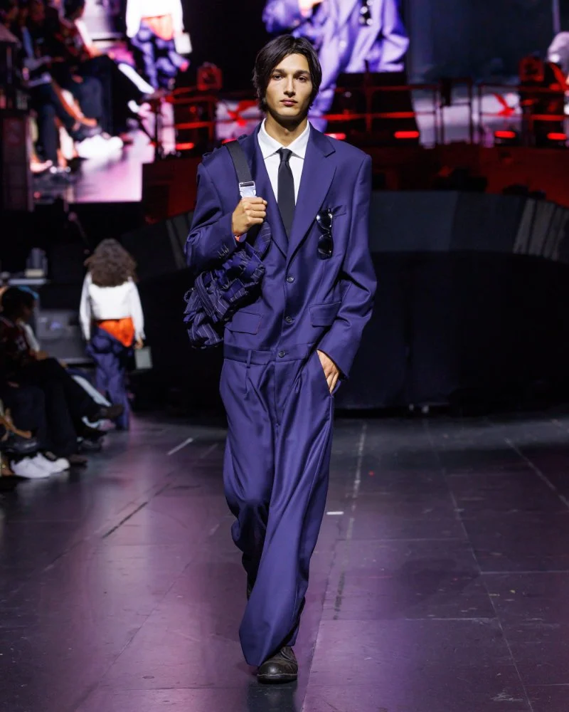Costume violet ample avec chemise blanche, cravate noire et sac porté à l’épaule, défilé AWGE printemps-été 2027.