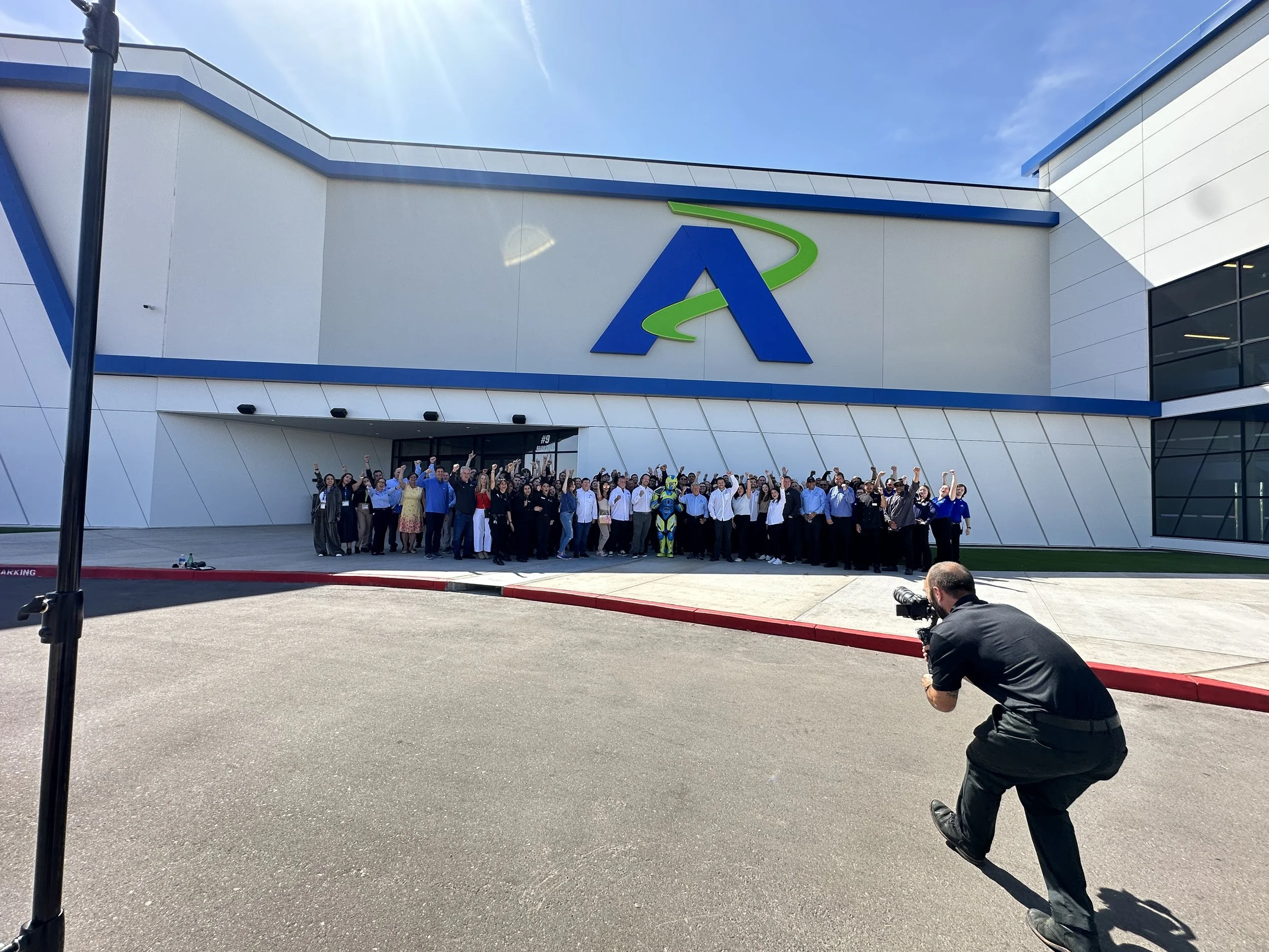 Group of people standing in front of an Andretti location, posing for photo. A photographer is taking picture from lower right. The sky is clear and sunny.