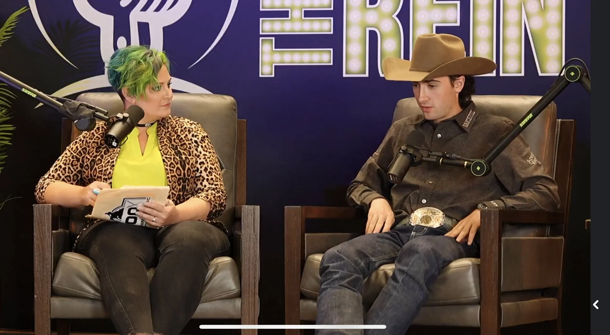 Chelsea Sutton with short, multicolored hair and Luca Fawearing a cowboy hat sit on chairs at a podcast or interview setup. She is dressed in a leopard print blazer and Luca Fappani in Western large belt buckle