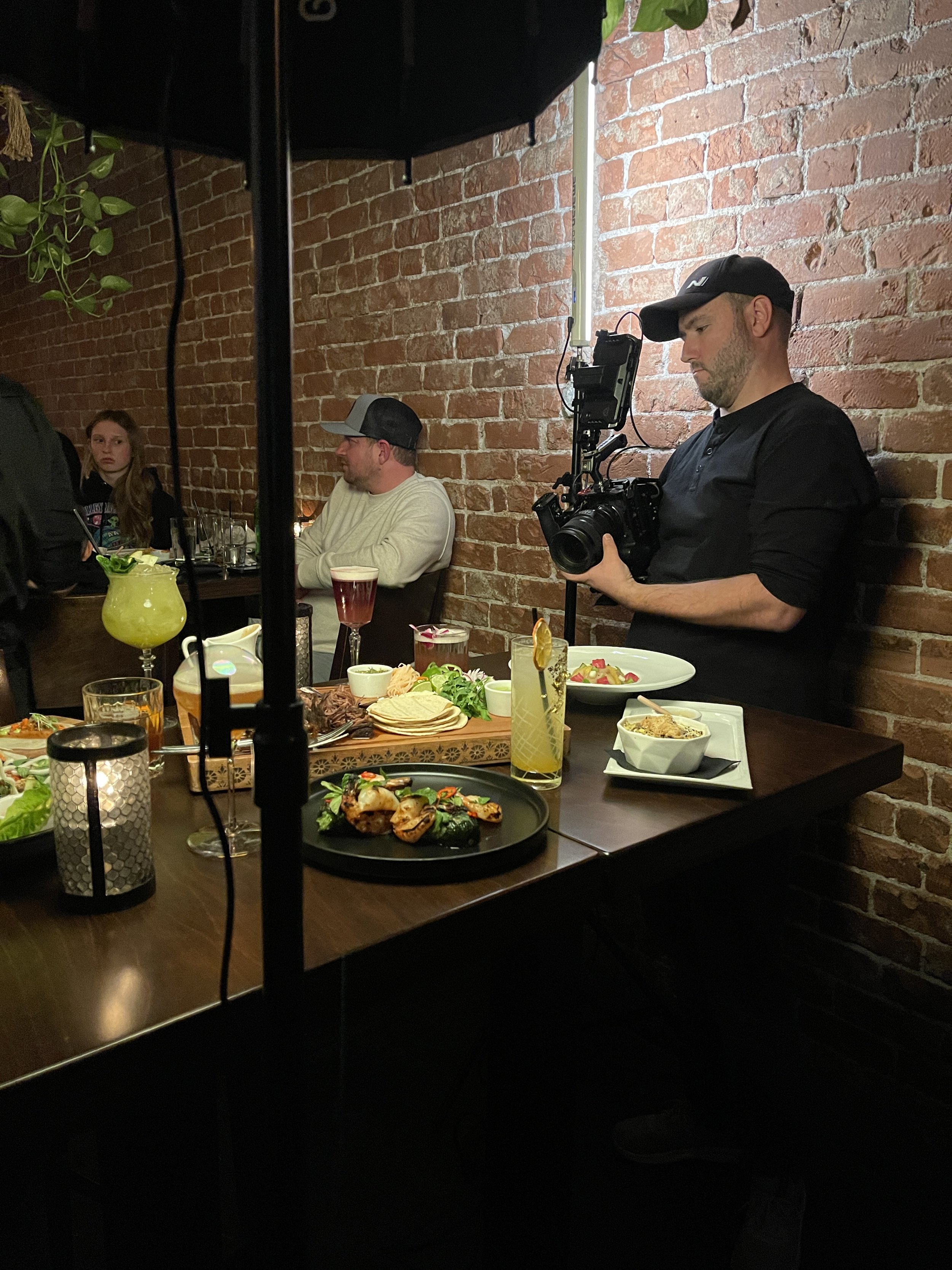 A man wearing a black cap and black shirt is standing in a restaurant kitchen, taking a photo with a camera. There are several colorful drinks and plates of food on a dark wood table, with a brick wall in the background and a girl sitting at a table in the background.