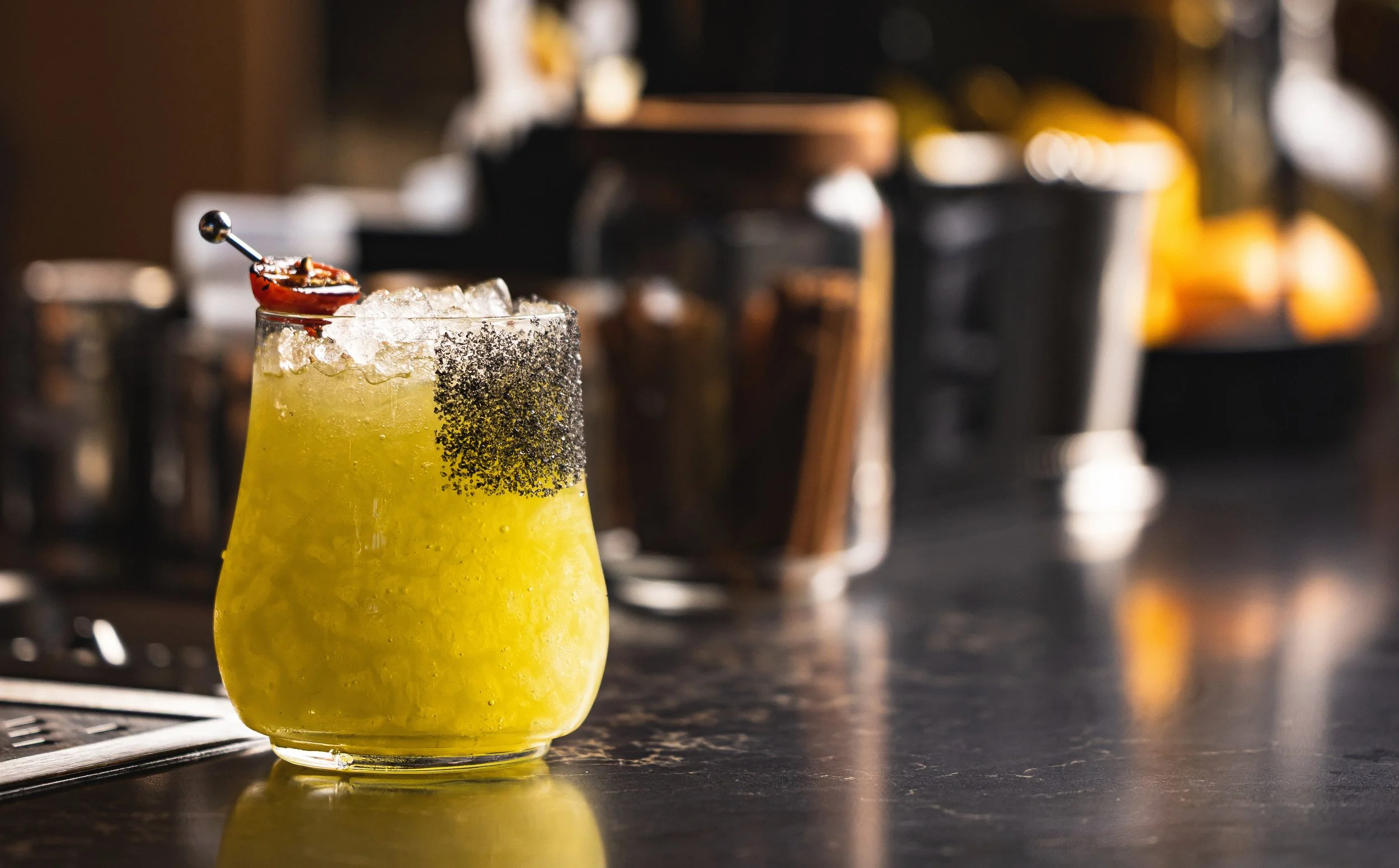 A yellow drink with ice in a clear glass, garnished with a cherry, in a bar or restaurant setting.