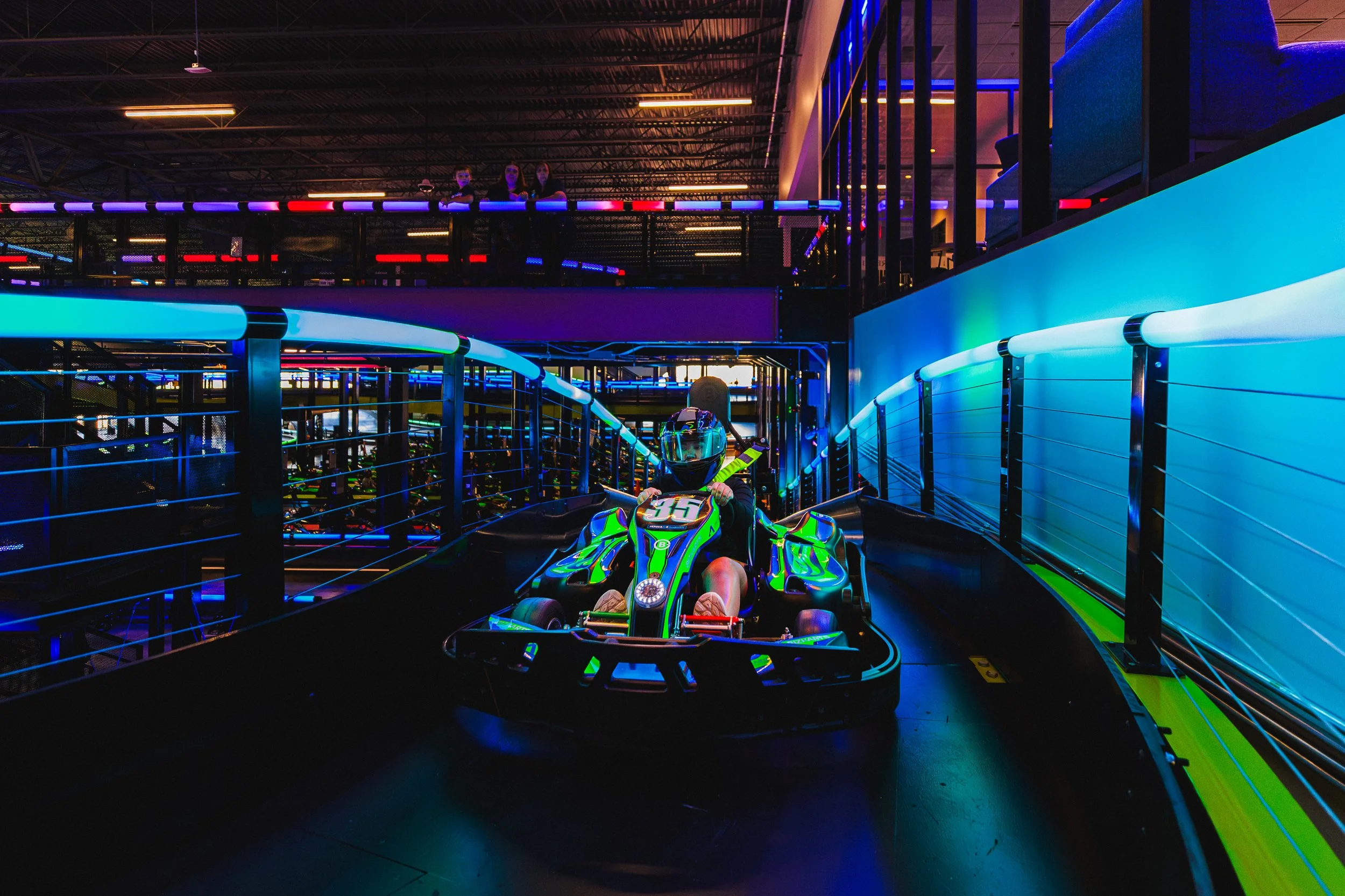 A person wearing a helmet and riding a neon green go-kart on an indoor track with blue and colorful neon lighting, with a balcony and three people watching from above.