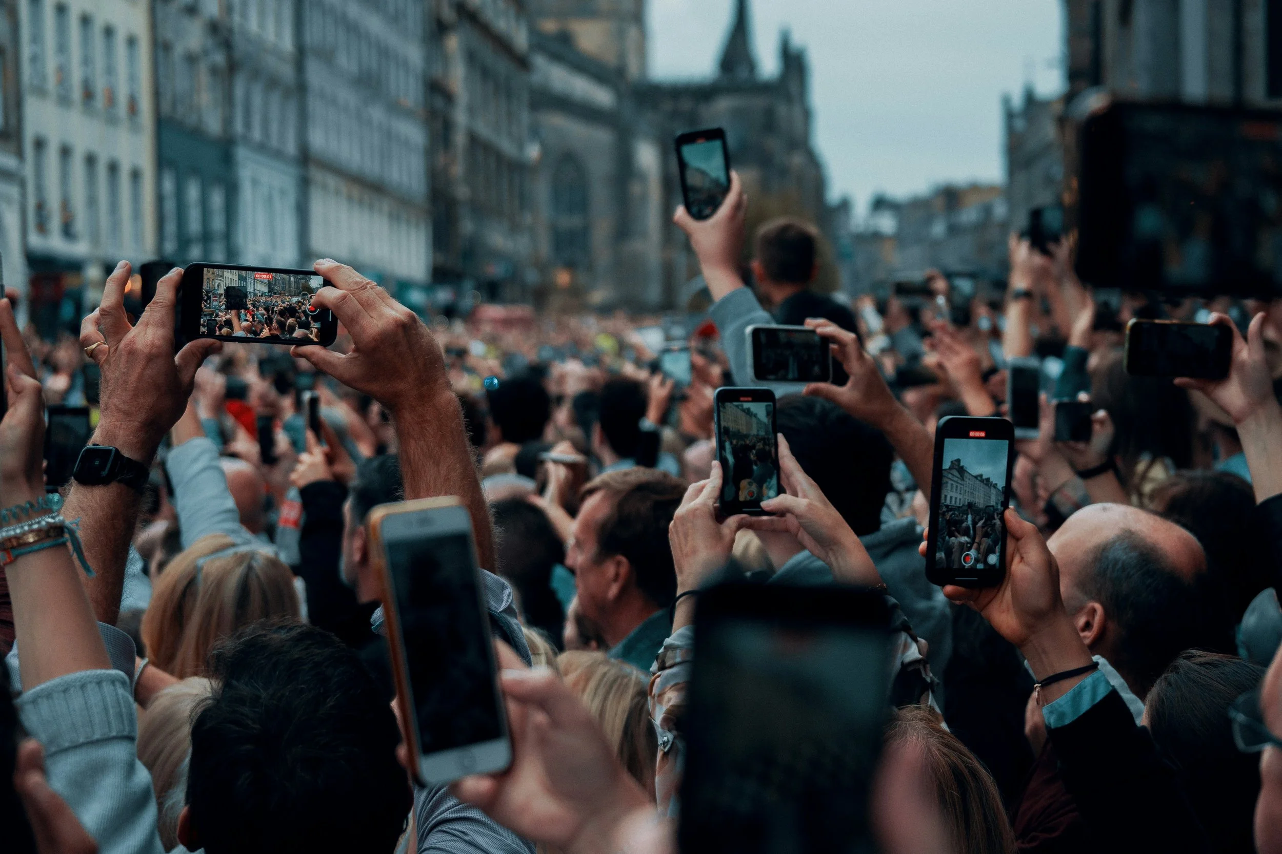 A large crowd gathered outdoors in an urban area, with many people holding up smartphones to record or take pictures of an event.
