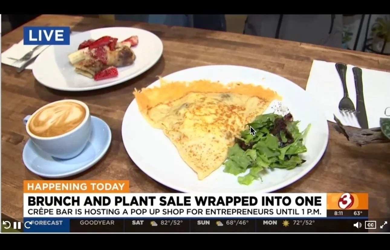 A press mention showing a table with a plate of crepes, a small green salad, a cup of coffee with latte art, and a smaller white plate with a dish topped with strawberries. A TV news banner at the bottom mentioning a brunch and plant sale event.