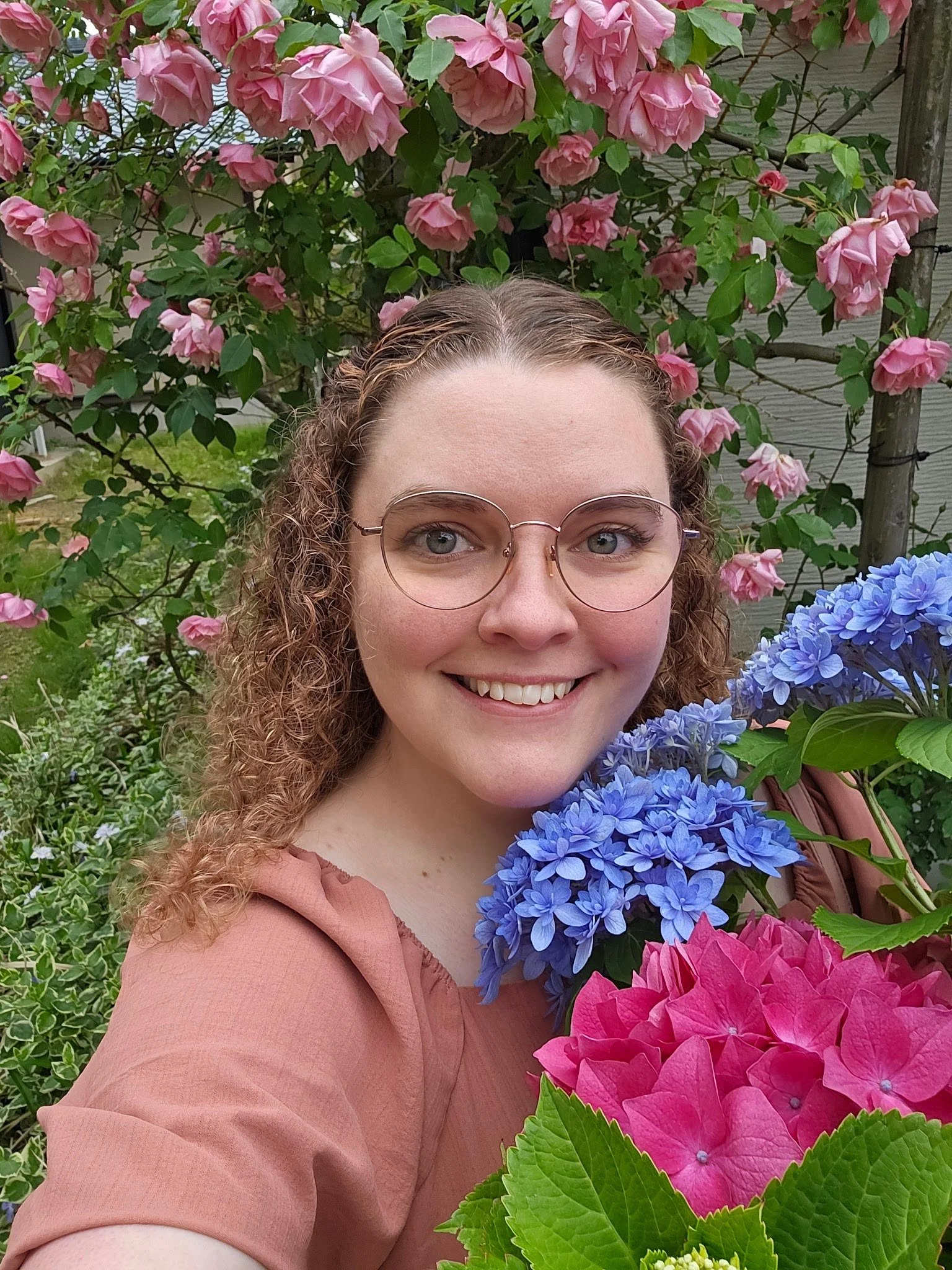 Casual self portrait of artist Abigail Zeigler. She has curly hair, glasses, and a terracotta colored dress. She is in front of pink climbing roses and holding a blue and pink hydrangea.
