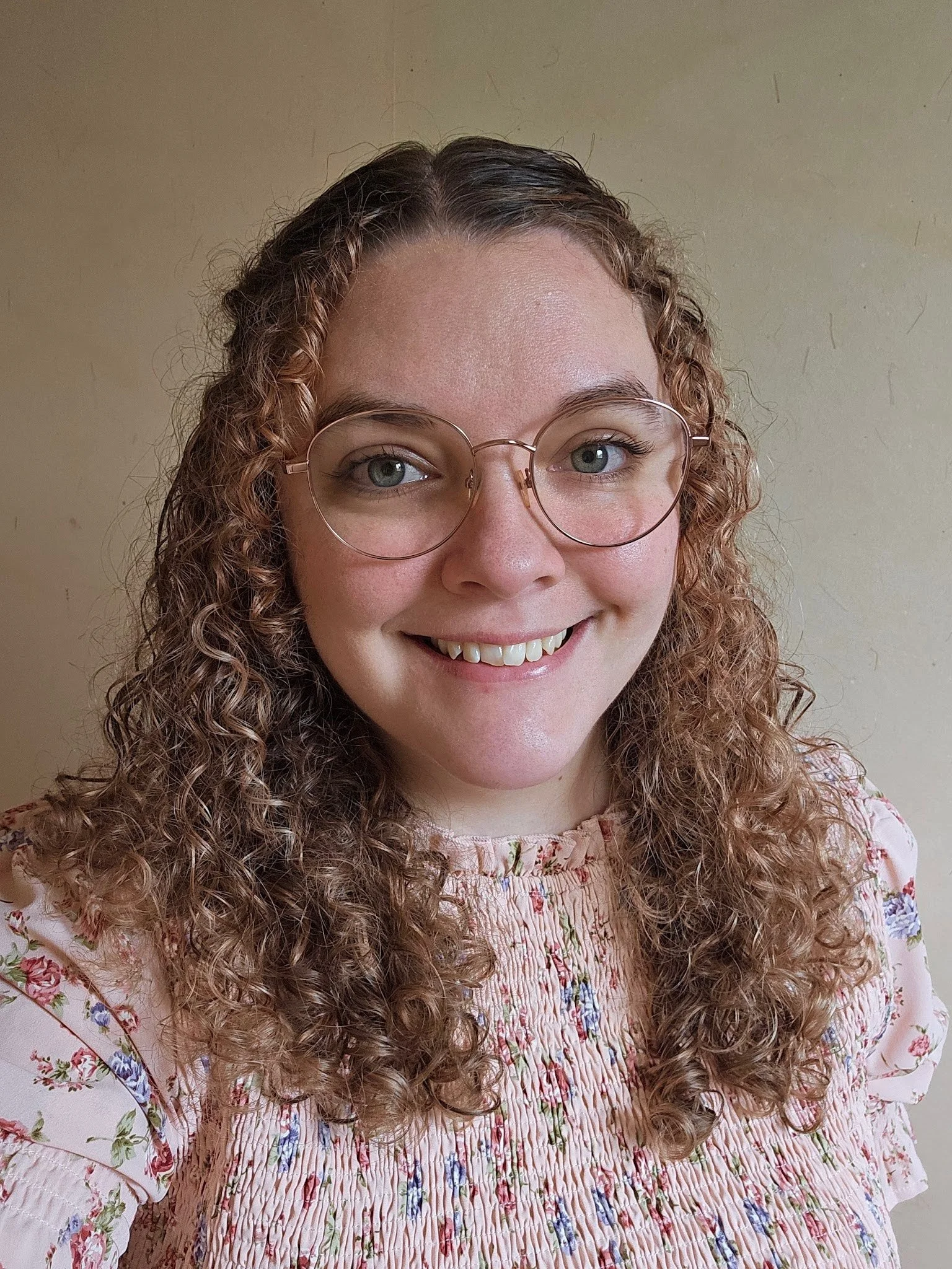Portrait of the artist Abigail Zeigler. She has curly hair, glasses, and a pink floral dress. She is smiling at the camera.