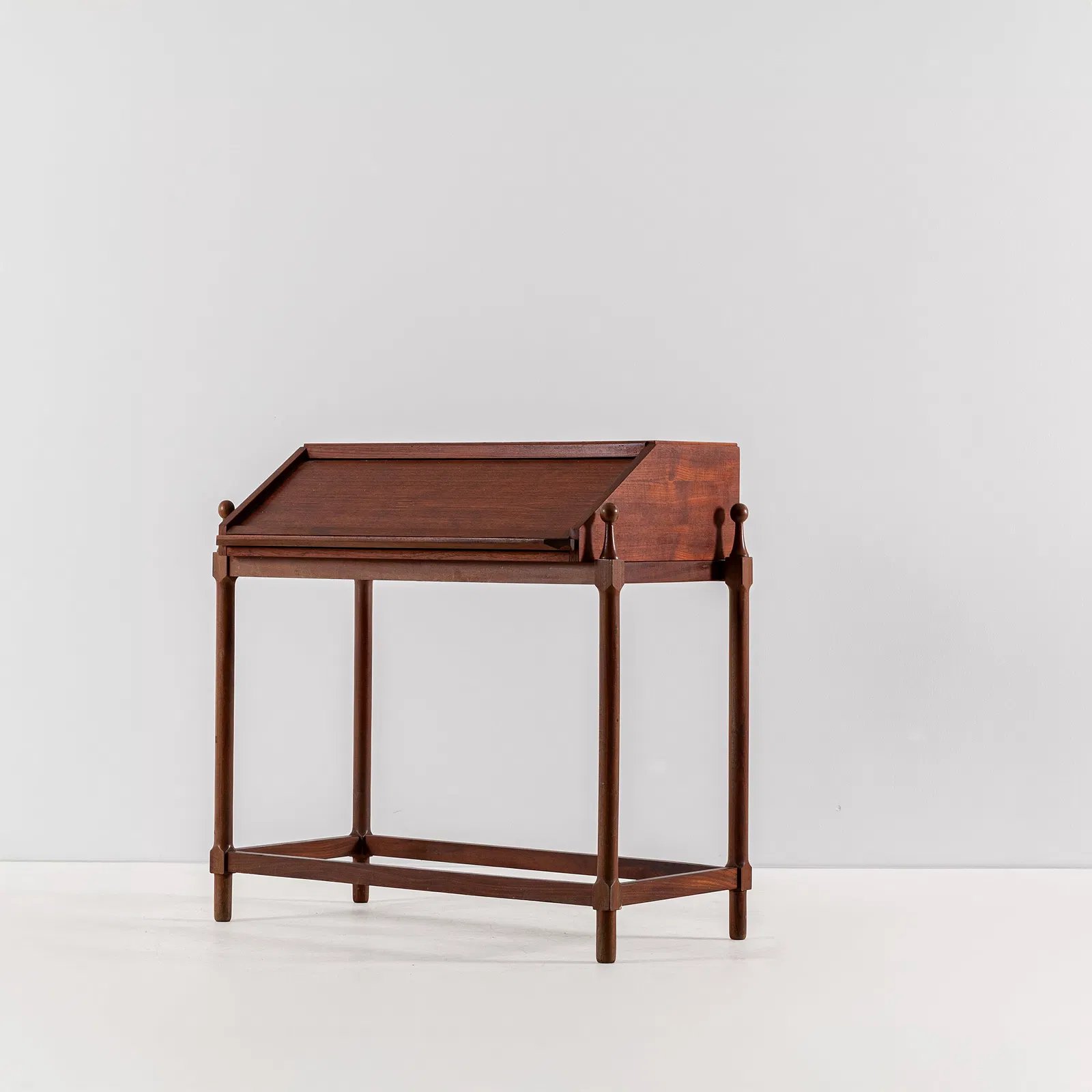 A vintage wooden writing desk with a sloped top and four legs, set against a plain white wall.